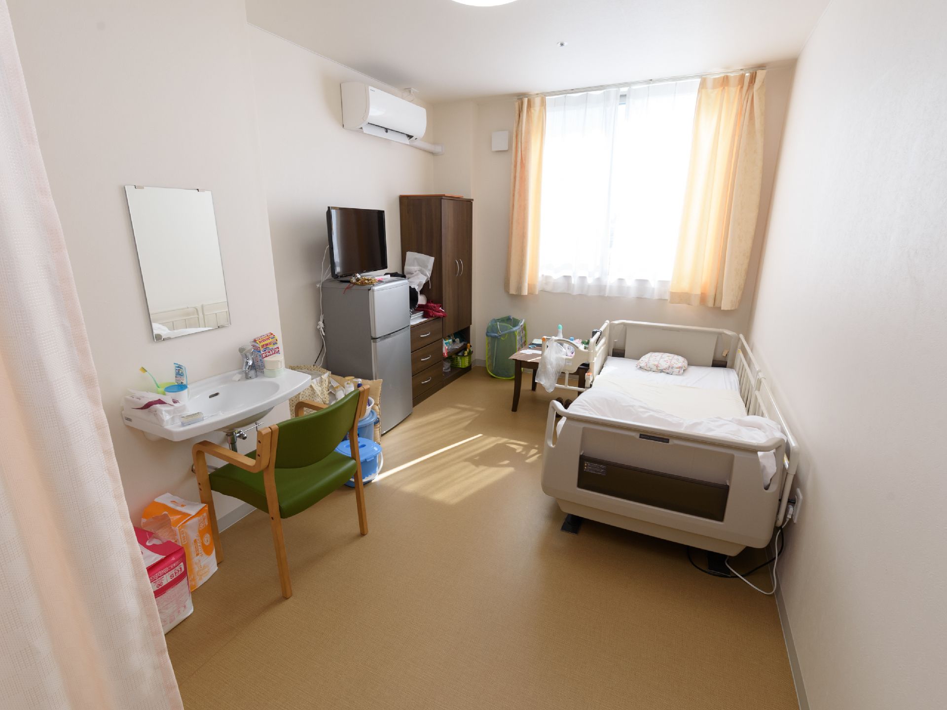 介護ベッドや収納家具、洗面台、冷蔵庫などが置かれている。洗面台周りには鏡や椅子もあり、冷蔵庫の上にテレビが乗っている。