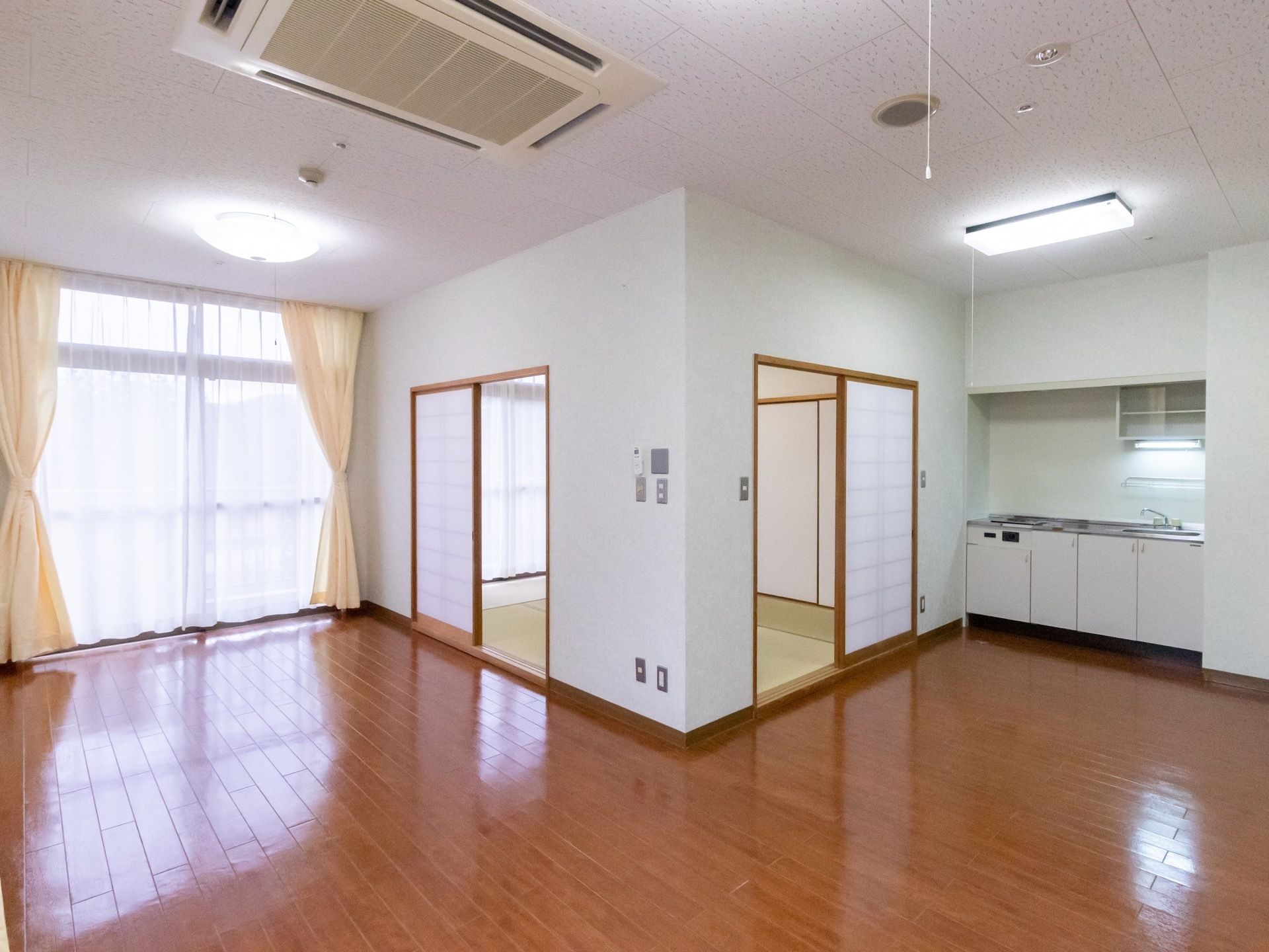 フローリング張りの居室で、中央奥に和室が備え付けられている。右奥にはキッチンがあり、コンロと流しが見えている。