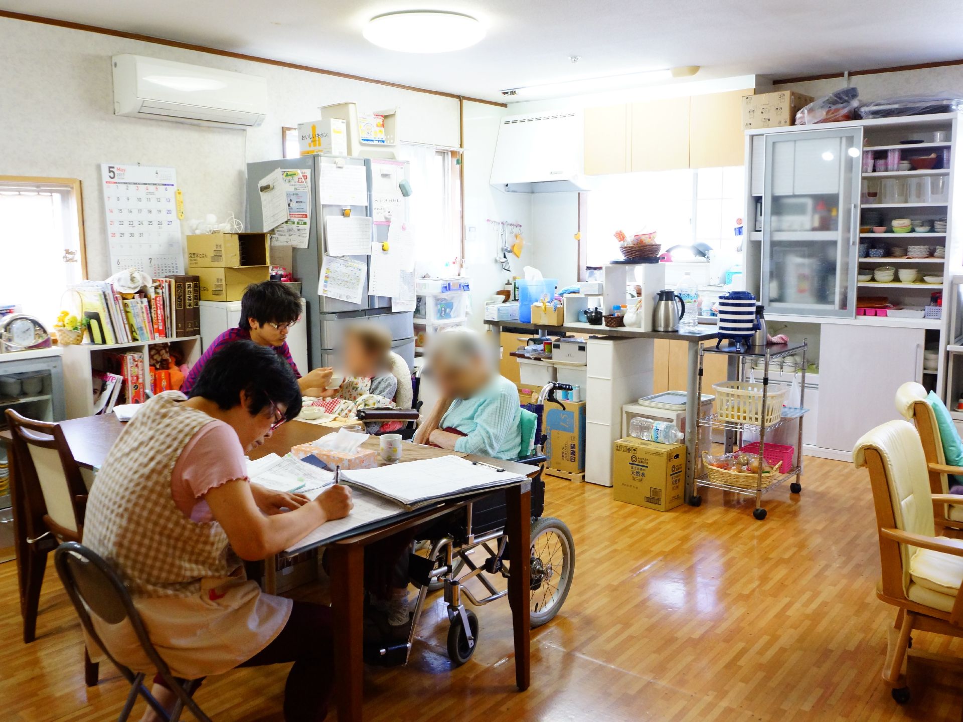 食堂で入居者様二人と職員が二人座っている。ひとりはスタッフと話をしている。スタッフの一人は書類に何か書き込んでいる。