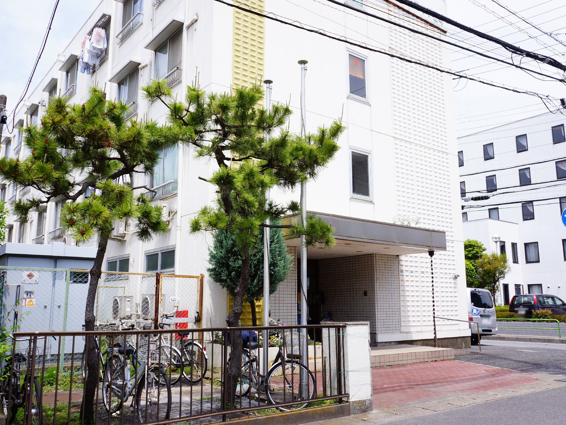 白いマンションのような作りの建物になっている。タイル調の玄関の手前にはフェンス越しに自転車が停められている。