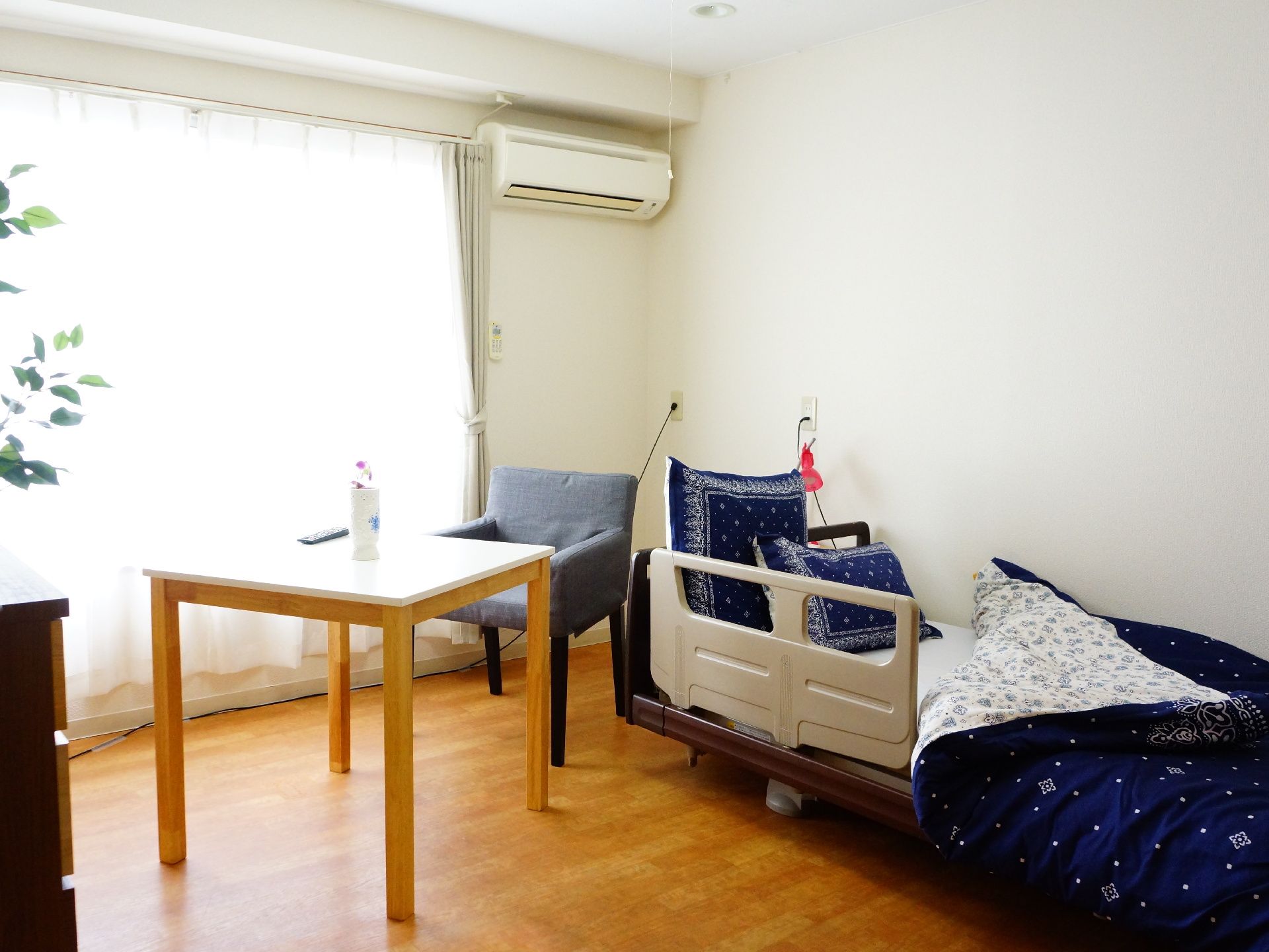 右上上部にエアコンが設置されとり大きな窓からは光が差し込んでいる。四角のテーブルと椅子がありベッドが右側にある。