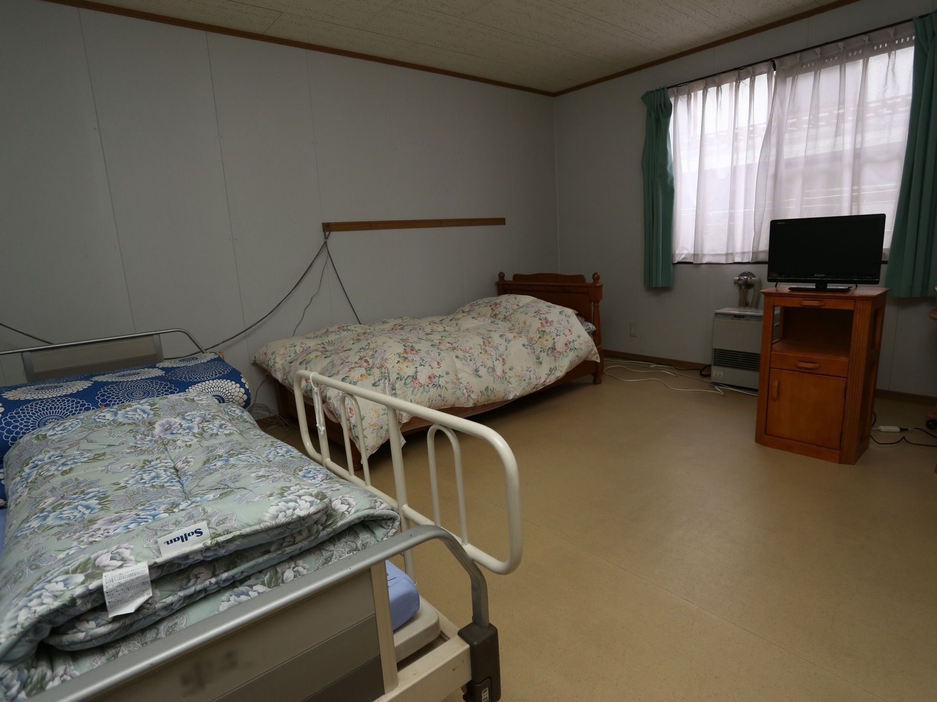 窓のある明るい室内にベッドが２台とテレビなどが設置されている。手前にあるベッドには白い手すりが付いている。