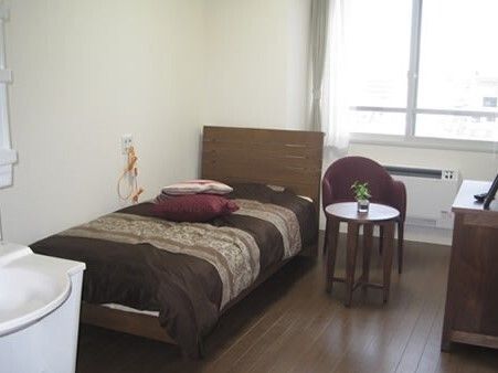ベッドサイドには緊急通報装置があり、ブラウン系の寝具がセットされたベッドが置かれ、円形の小さなテーブルには植物が飾られている。