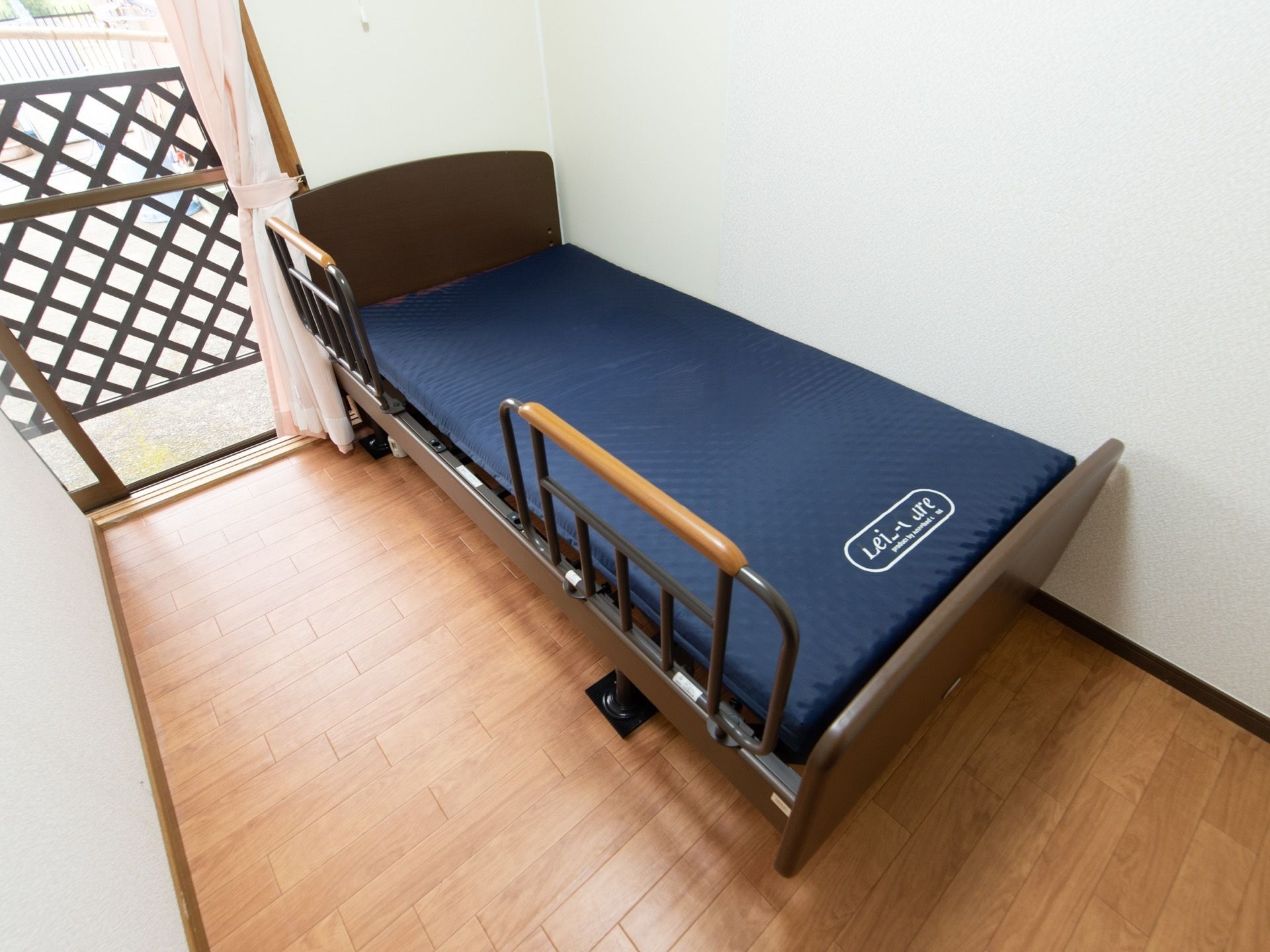 木のフレームのベッドがあり、青いマットレスが敷いてある。ベッド柵には、持ちやすいように厚くなった部分がある。