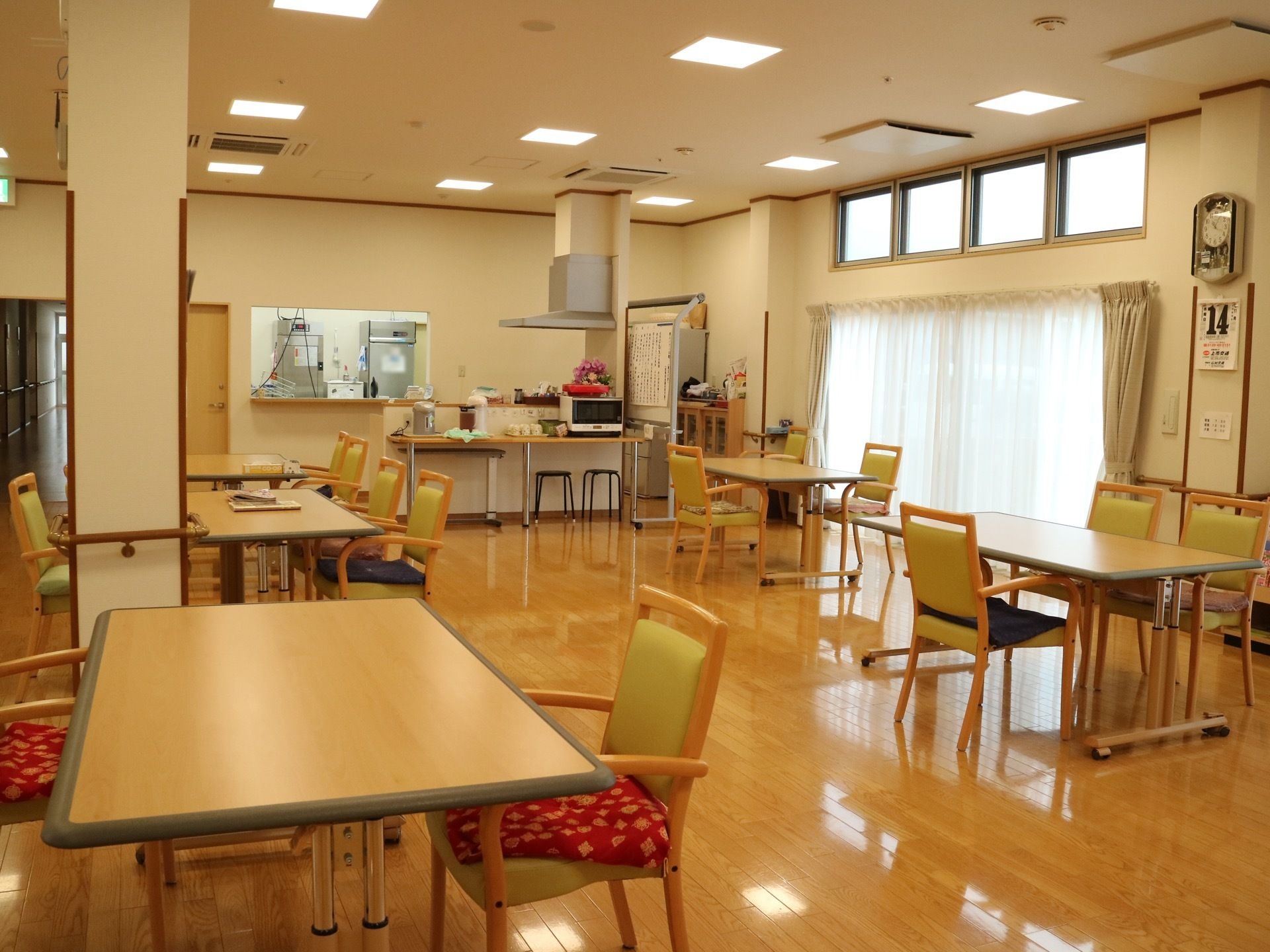 大きな窓がある食堂である。時計やカレンダーが壁に掛けられている。椅子にはさまざまなクッションが使われている。