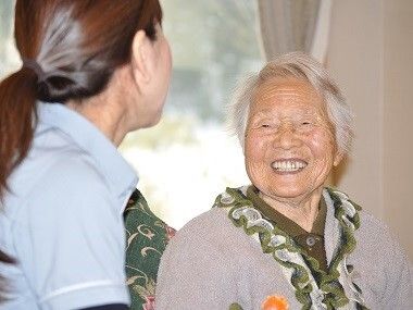 ピンクのポロシャツを着た若い女性スタッフと白髪の高齢女性がテーブルを囲んで話している。