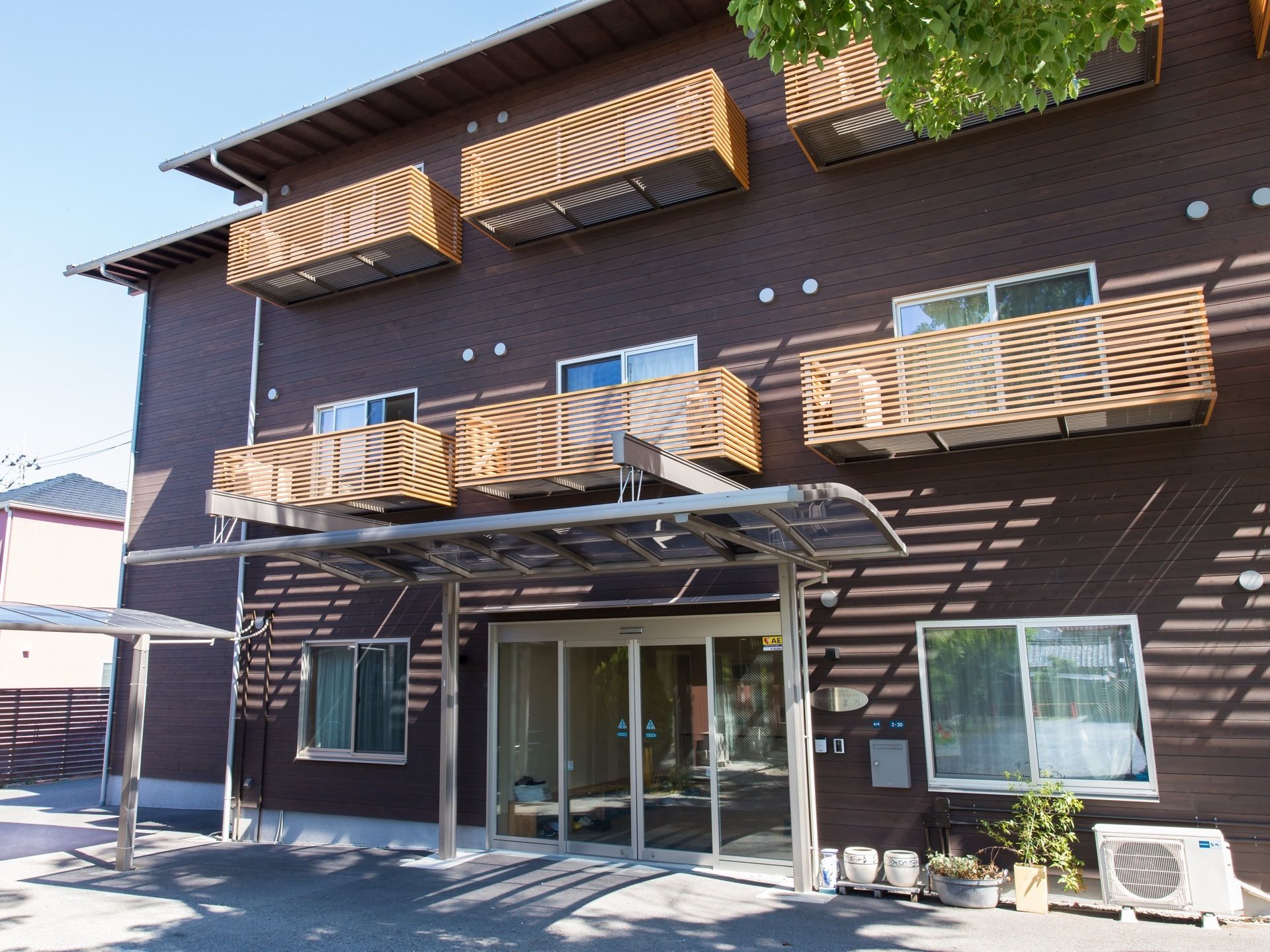 施設は木造３階建てである。外壁は茶色に塗られている。２階、3階にはバルコニーが設置されている。玄関脇に草木の鉢が置かれている。