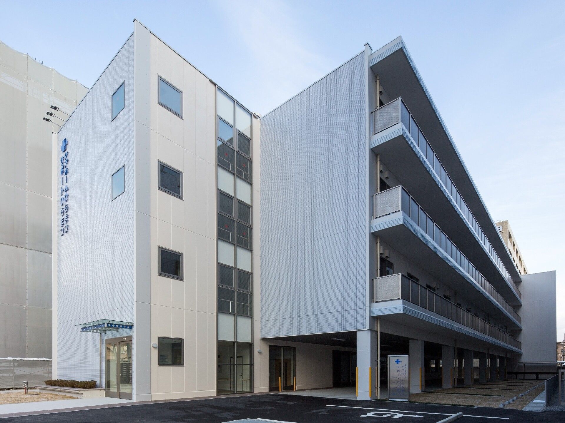4階建ての建物の各フロアにベランダがあり、敷地内には駐車場があり、周りにも建物が建っている。