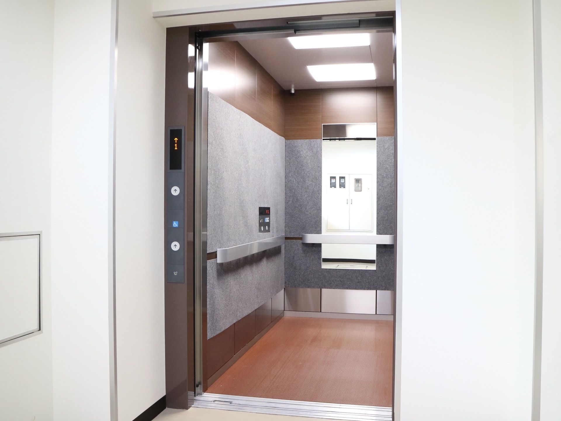 奥行きのある広々としたエレベーターで、中には手すりや鏡、操作パネルがある。天井には照明が２つ取り付けられている。