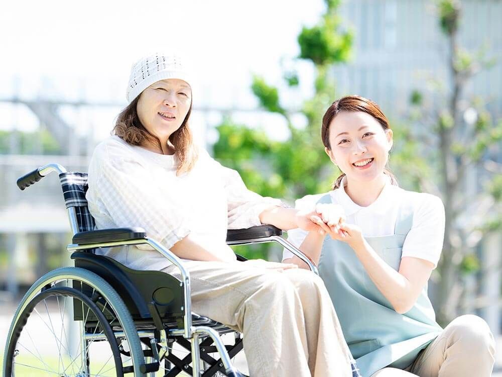 車いすに乗っている女性とその人の手を握りながらこちらを向いて笑顔を見せるスタッフ