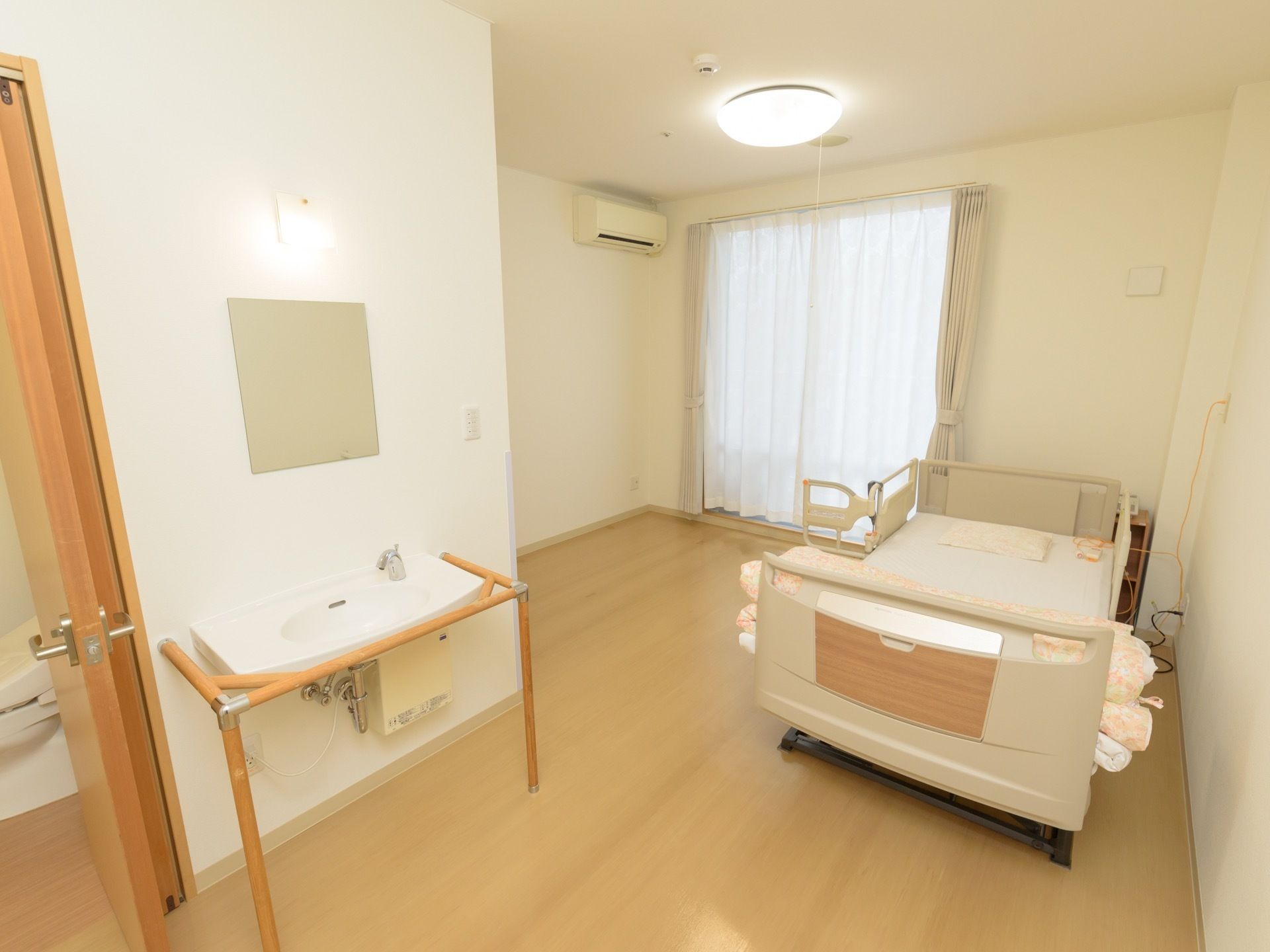 手洗い場の隣にはトイレが設置されており、木目の開き戸がついている。電動ベッドが居室の隅にあり、緊急通報装置がベッドの上に置いてある。