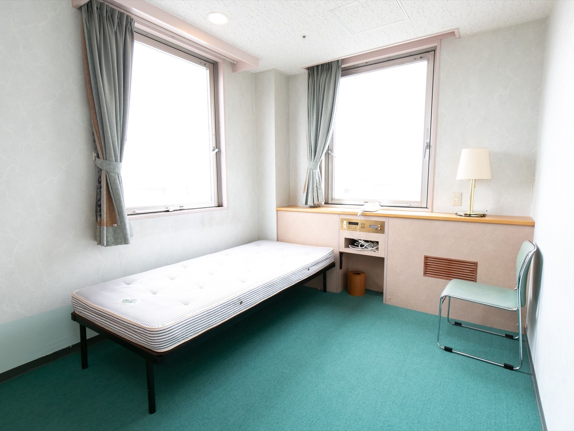 緑色のカーペットが敷かれた居室がある。窓が２つ付いている。壁の一部が棚になっており、照明器具がある。マットレスの置かれているベッドがある。
