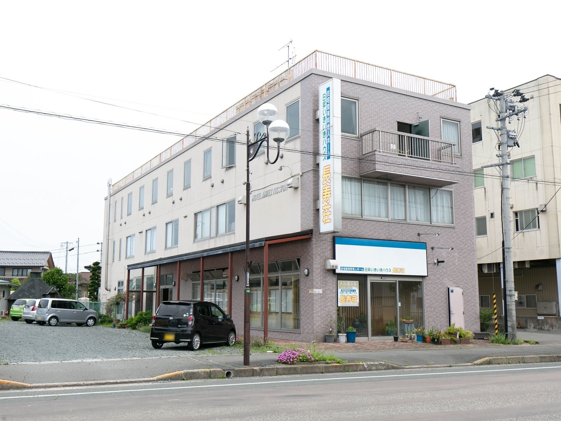 道路に面した位置に、３階建ての建物がある。大きな看板が掛けられている。敷地内には広い駐車場があり、車が停められている。