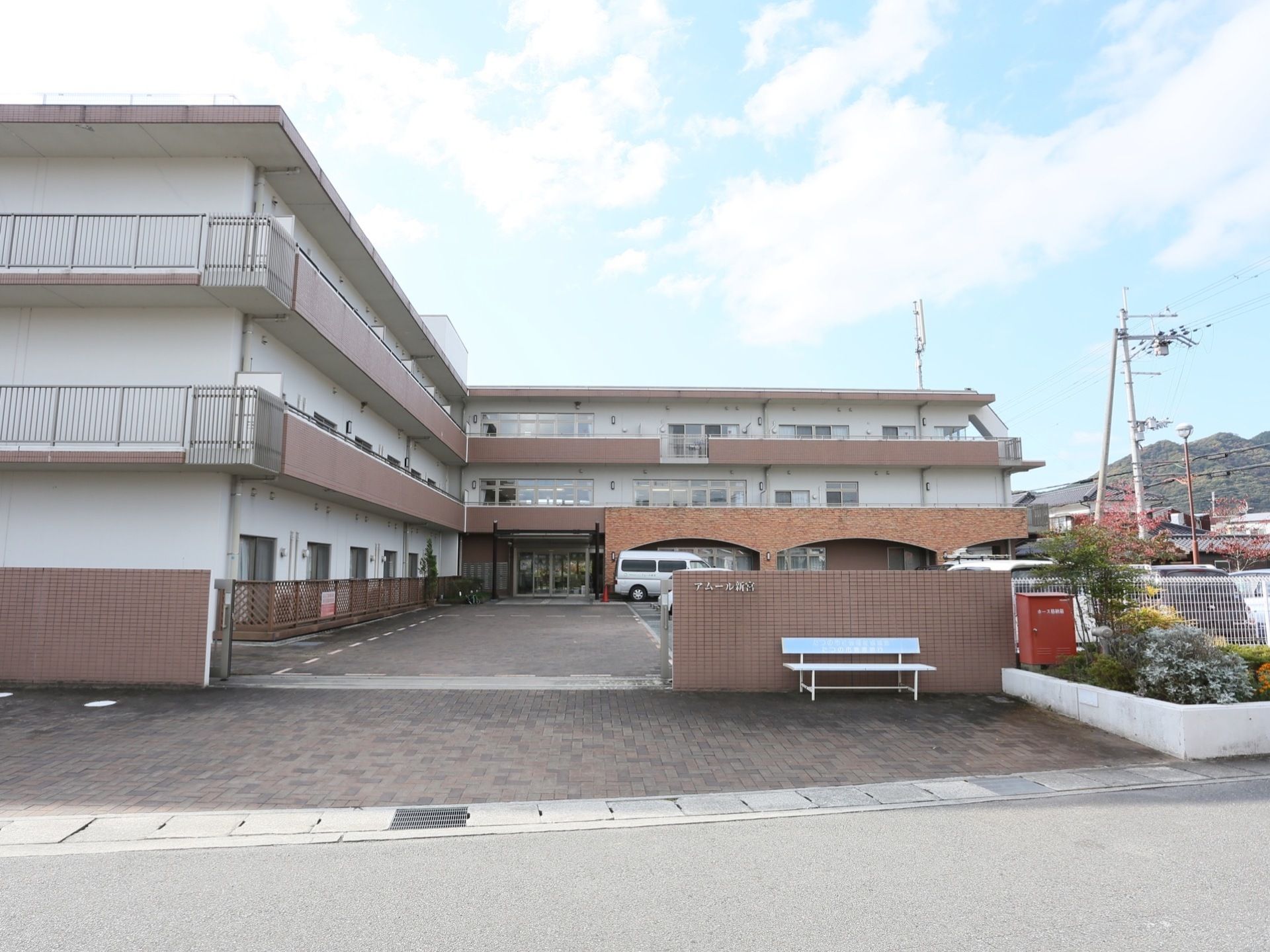 施設の前を通る道路から見た建物の外観である。Ｌ字型の建物は３階建てで、白色と茶色を基調としており、門の前にはベンチが置かれている。