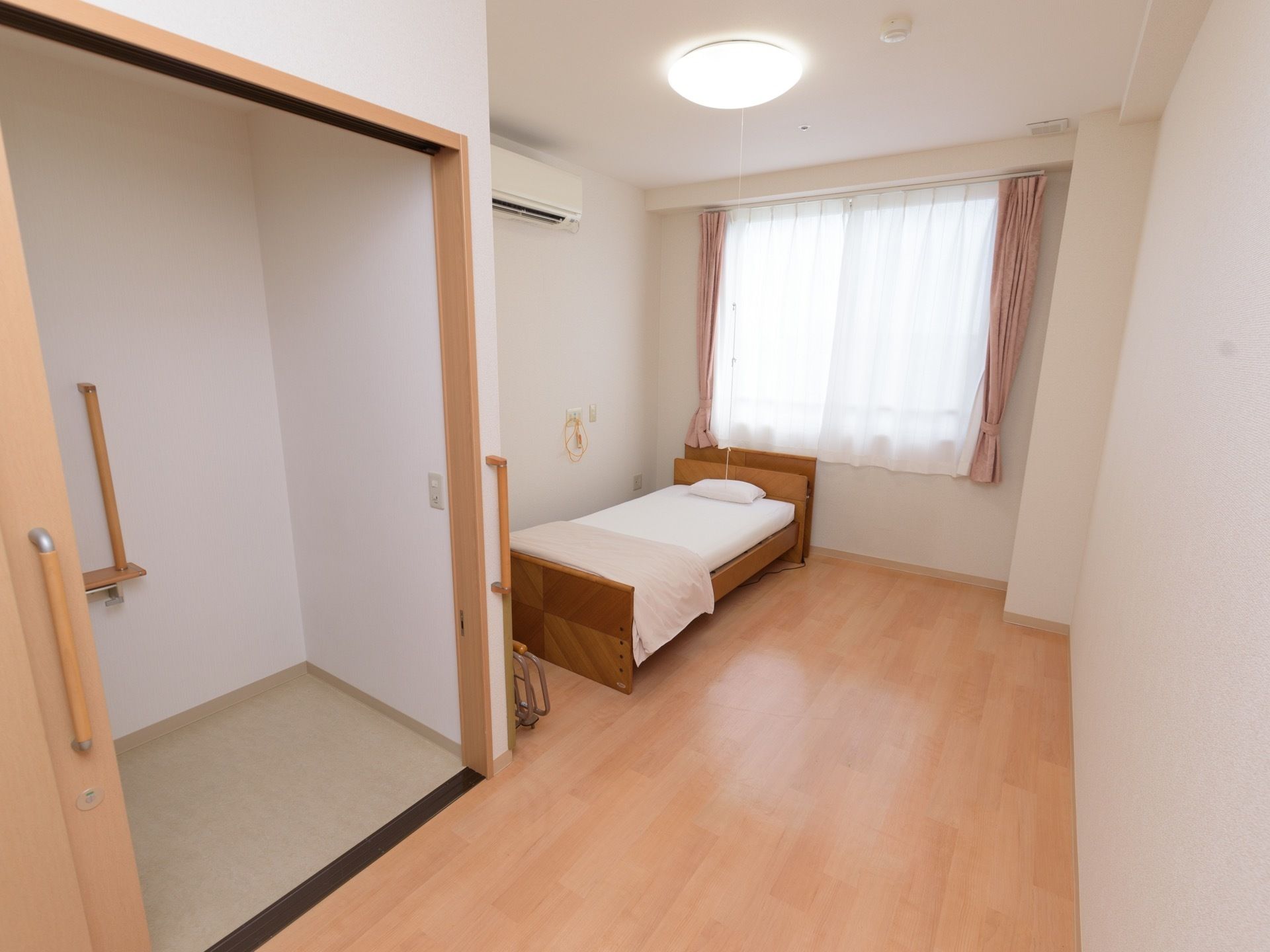 窓にはカーテンがかけられ、ベッドが１台置かれている。また、エアコンが設置されており、天井のシーリングライトからは紐が垂れている。