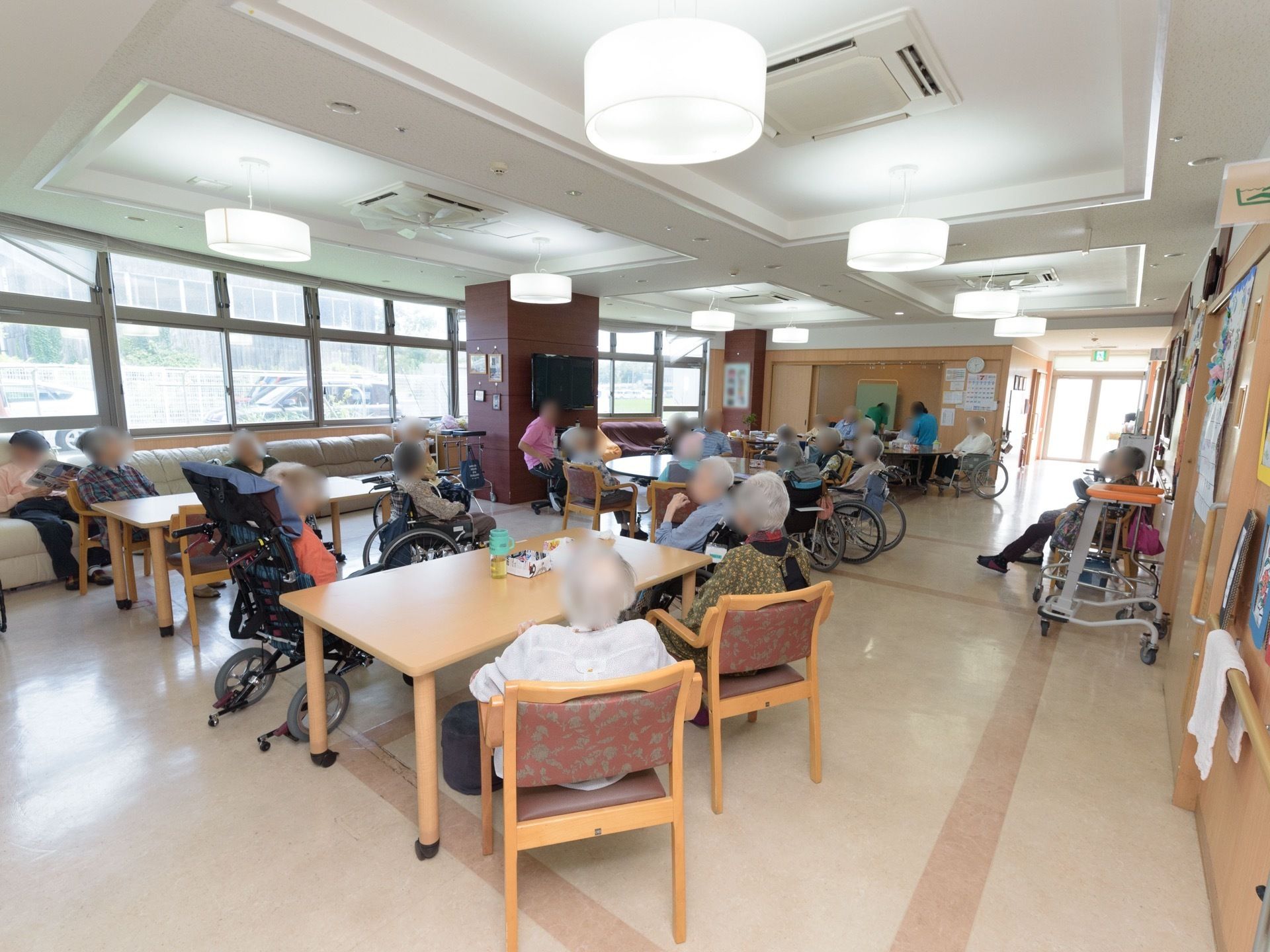 丸や四角のテーブルが複数置かれている。窓側には大きなソファが置かれており、入居者様が座っている様子である。