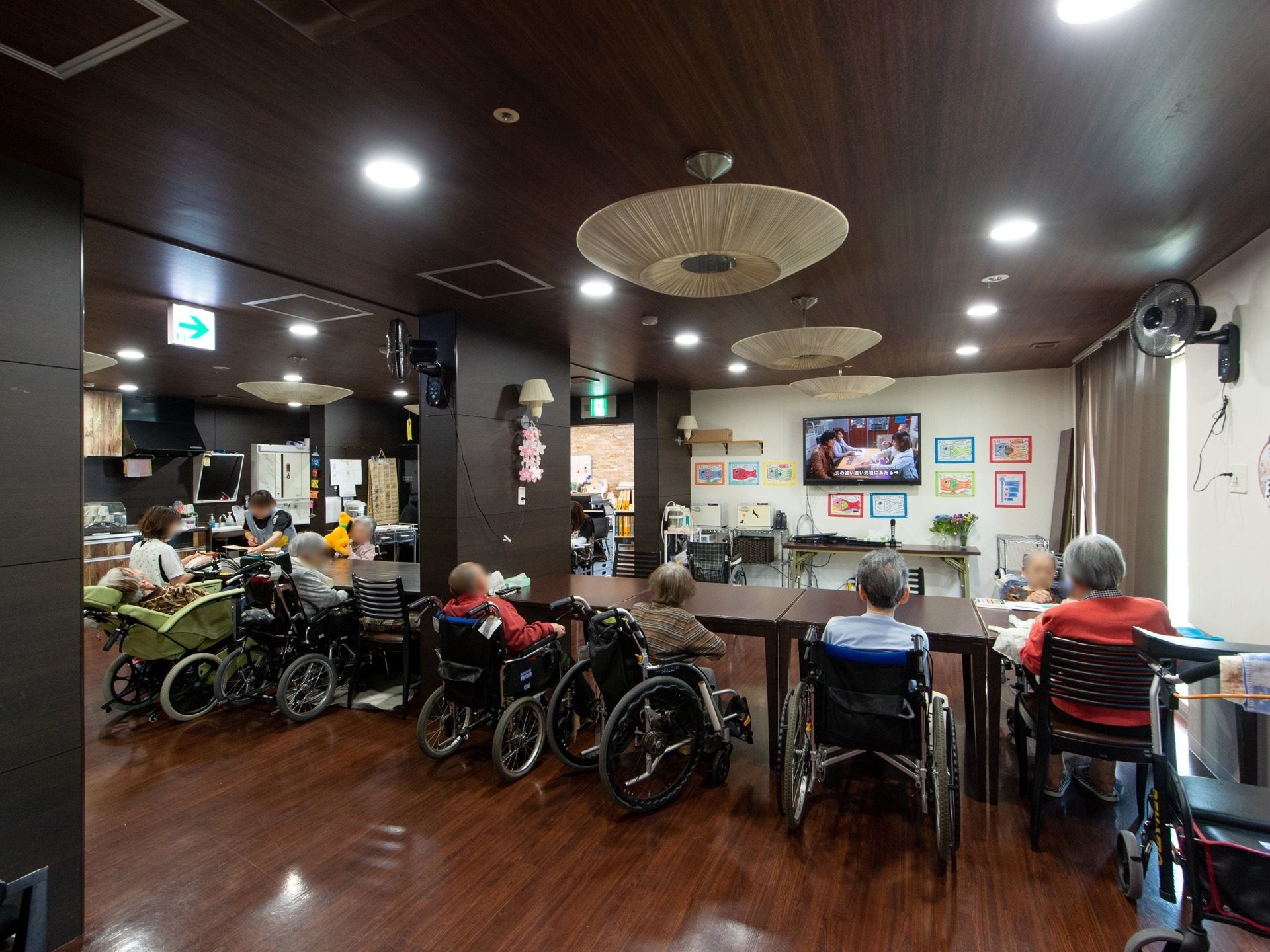 こげ茶色のテーブルが横に長く置かれており、入居者様が座ってテレビをみている。車椅子で席についている人もいる。