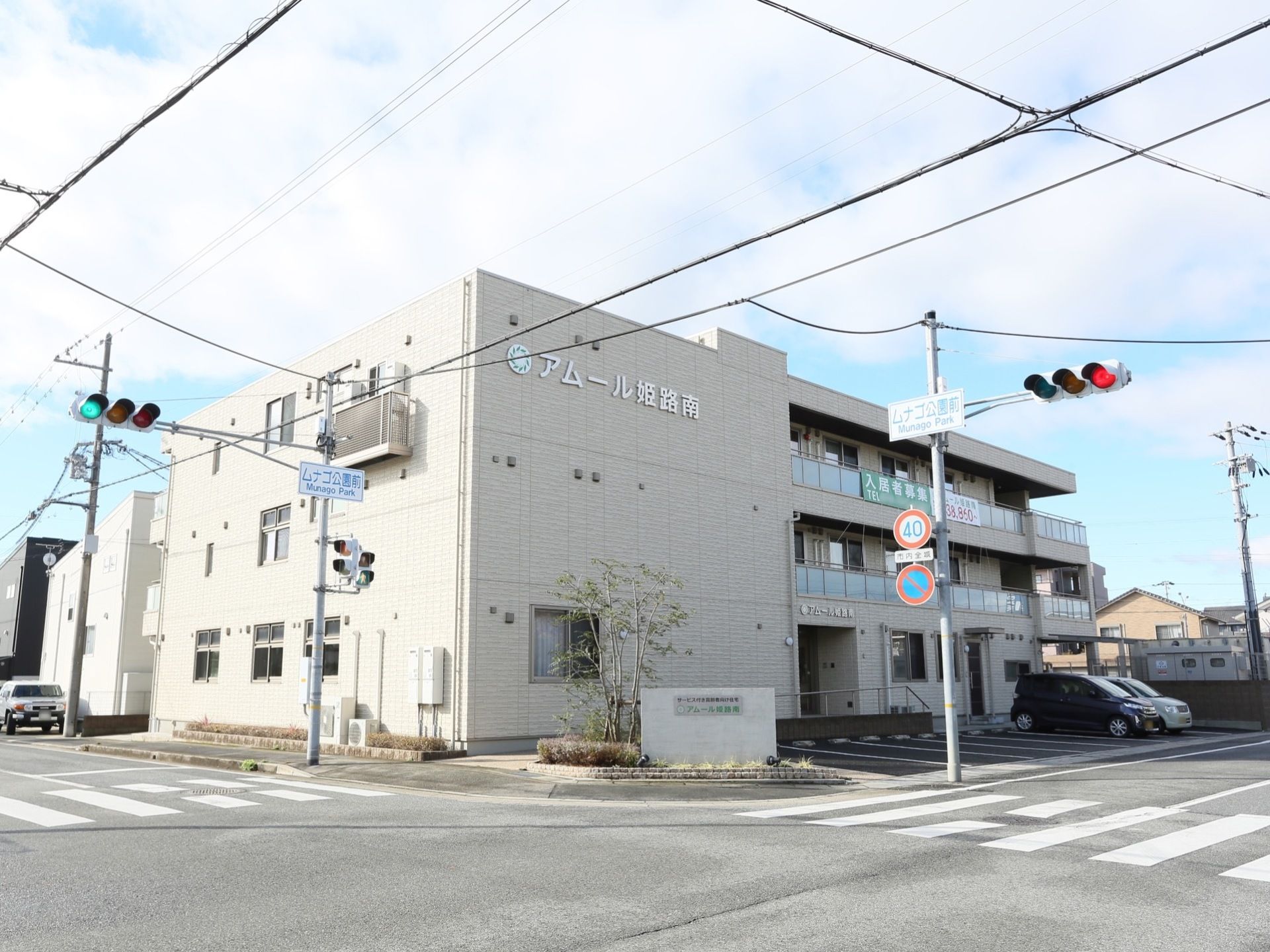 ３階建ての建物が交差点の角地に建っている。道路に面して駐車場があり、車が２台停められている。敷地の角に植え込みがある。