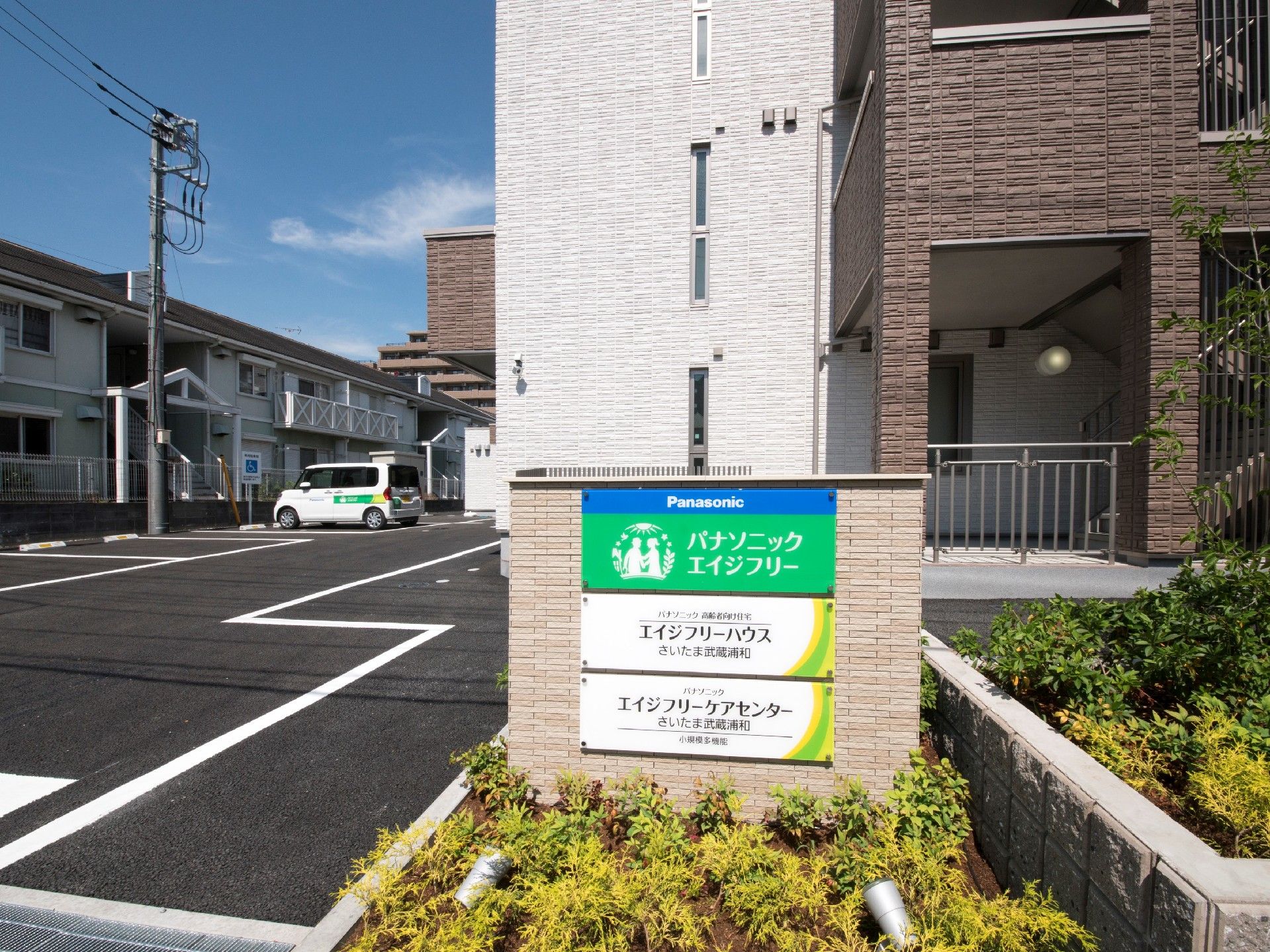 住宅や併設事業所の名前が書かれた門のそばには木が植えられ、駐車場や建物が奥に見える。