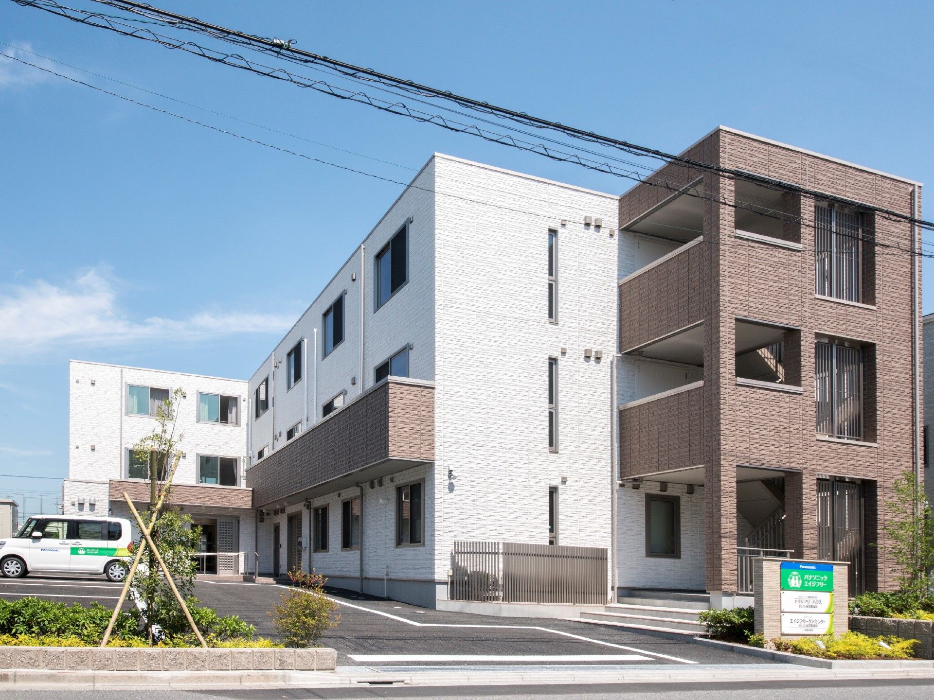 白とブラウンの3階建ての建物の周りには緑も多く、玄関には屋根がある。