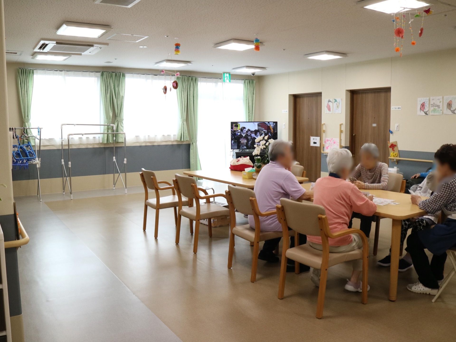 複数の窓が設けられている明るいフロアで、テレビや物干し竿が設置されている。入居者様が、思い思いの時間を過ごしている。