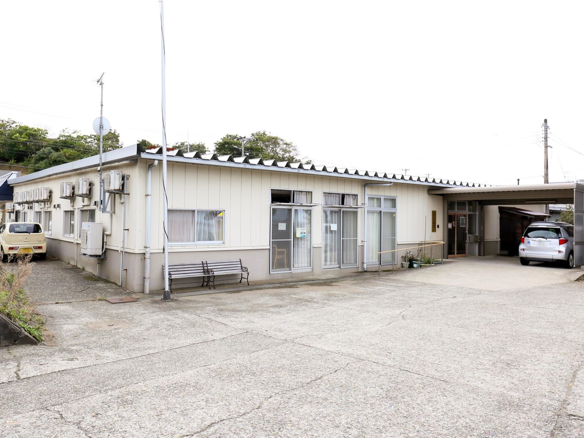 白色の平屋建ての施設の建物で、玄関前には車寄せが設けられている。建物に沿って2人掛けの屋外用のベンチが2台置かれている。