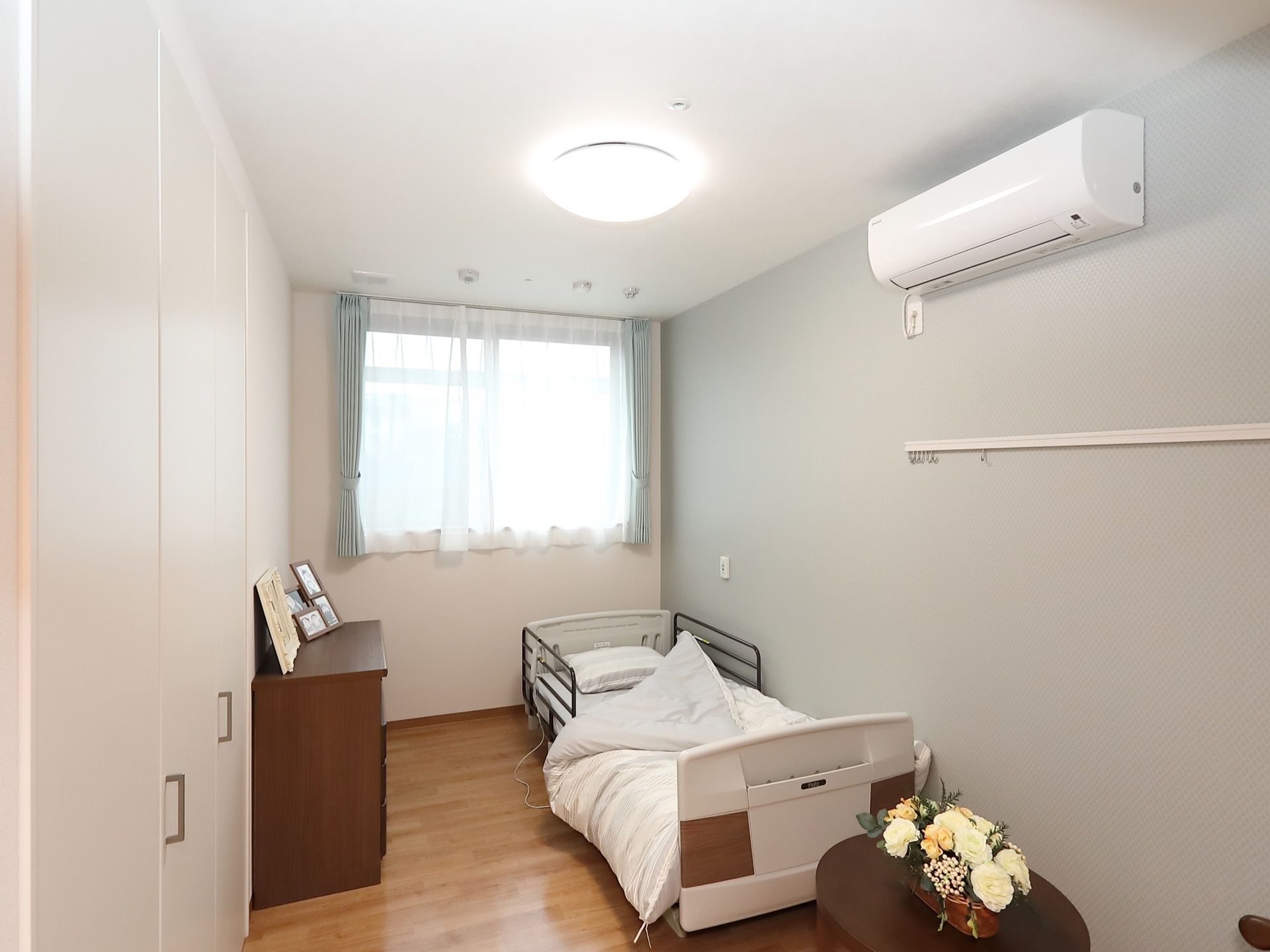 窓がある部屋にベッドが置いてある。右側には淡い灰色の壁紙が施されており、高い位置にエアコンを設置。