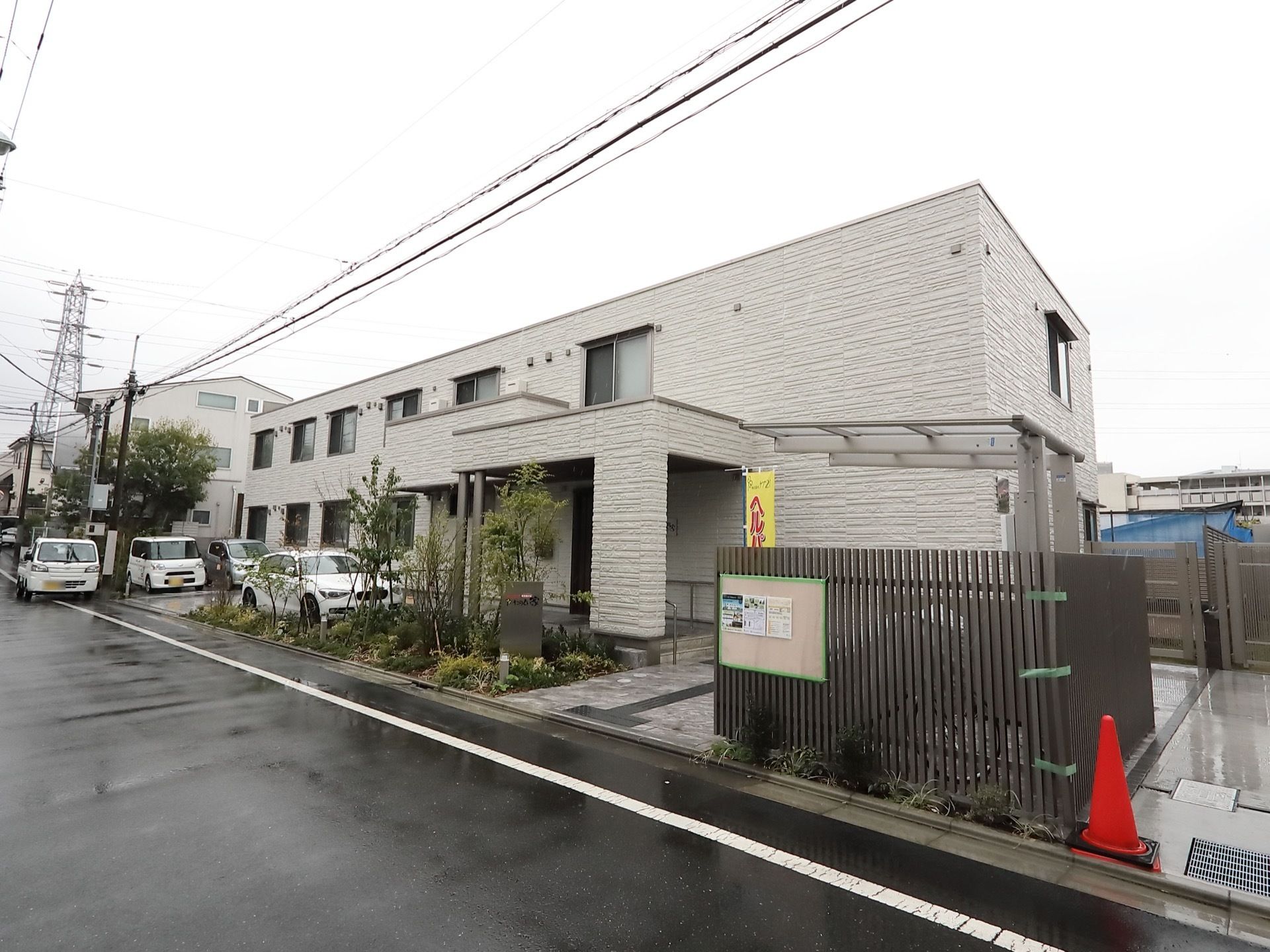白い外壁が施された横幅の広い２階建ての施設が車道に面して建っている。玄関の前に植栽が施されているスペースがある。