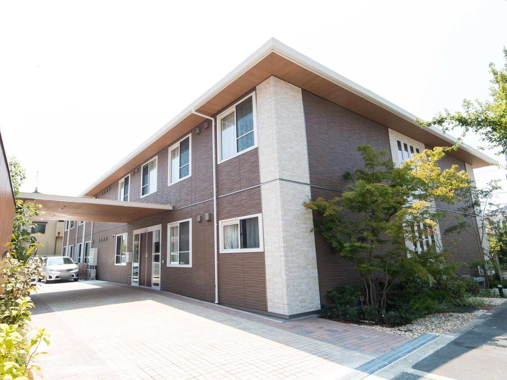 玄関前には屋根があり、奥に車が数台停まってる。建物の周囲や敷地内には、大きな木や植物が複数植えられている。