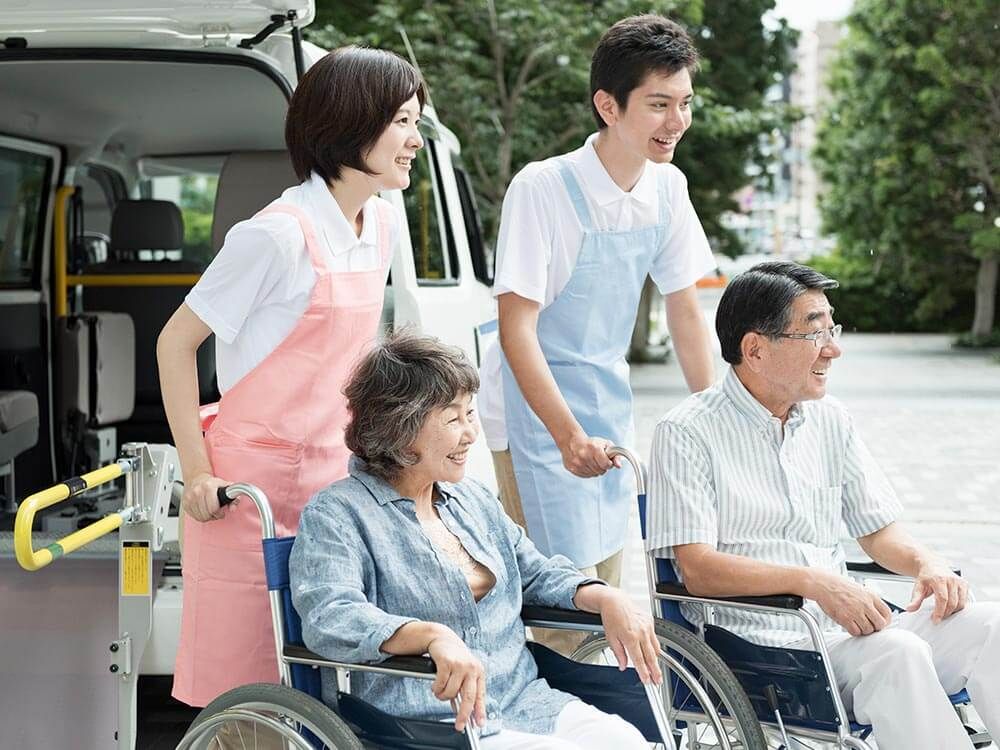 車椅子に乗った高齢の男女に付き添うヘルパー