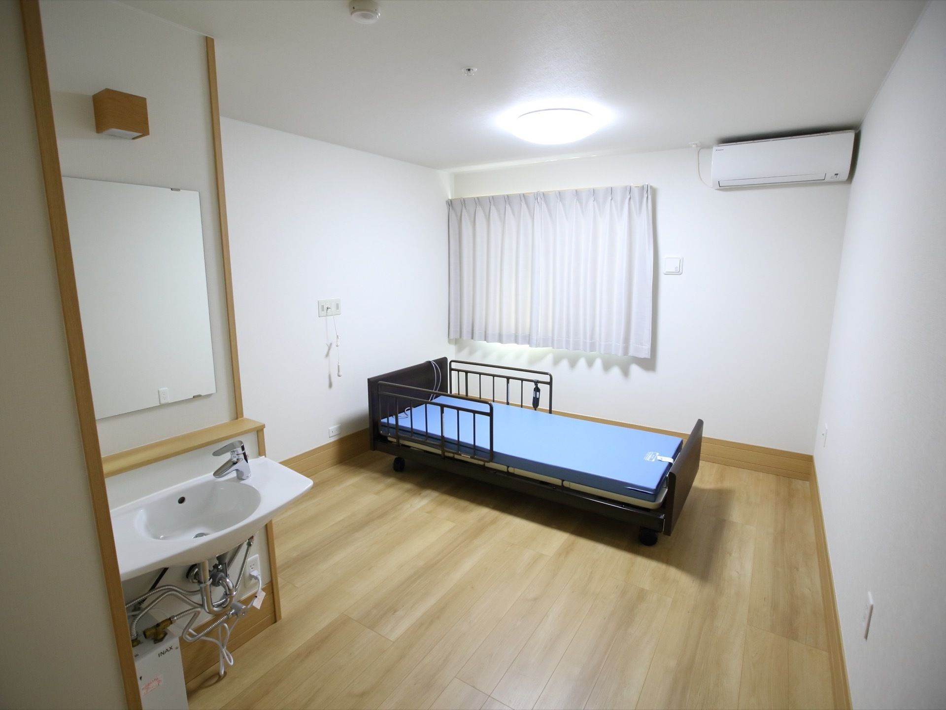 居室の入口付近に洗面台がある。窓の近くには、転落防止柵がついたベッドがあり、窓の右上にはエアコンがかかっている。