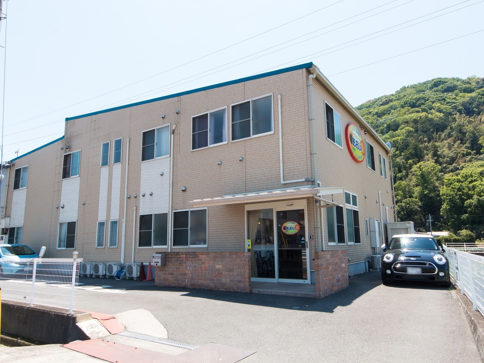 2階建ての大きな建物があり、その脇に黒い乗用車が留められている。建物の奥には一軒家や森も見えている。