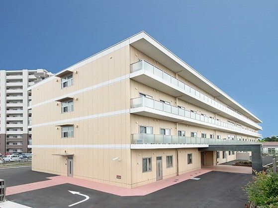 4階建ての建物はベージュ系の外壁で玄関には車寄せがあり、周りには緑も植えられている。