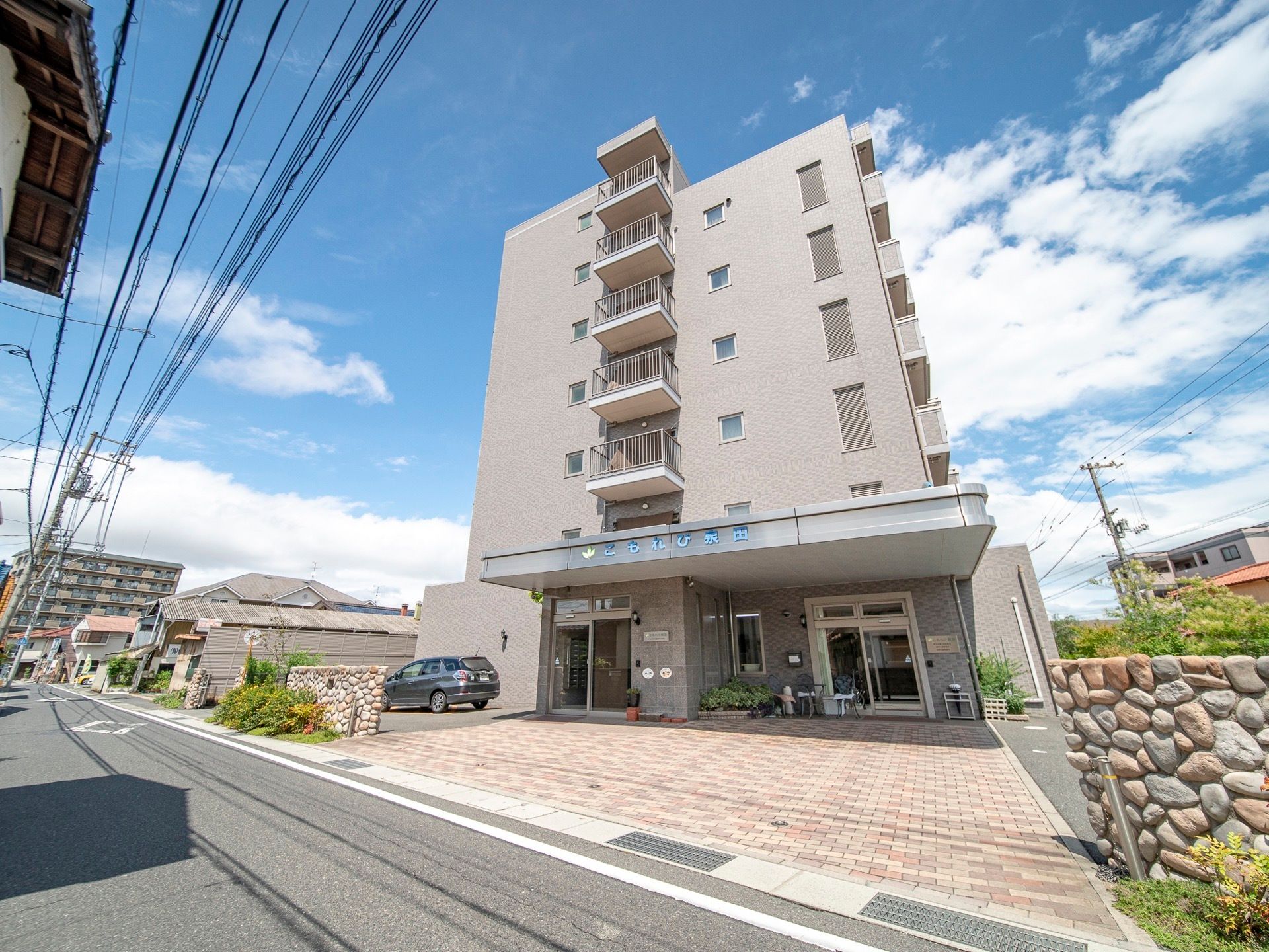 地上７階建ての施設の前に自動車が１台駐車してある。道路と敷地との間に段差が少なく、間口も広くなっている。