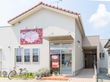 白い平屋建ての建物がある。外壁に施設名称の入った赤い看板が掲示されている。植栽スペースの前に１台の自転車が停められている。