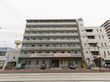 建物の前が駐車場になっており、数台の車が停まっている。２階バルコニーに入居者募集の広告がされている。