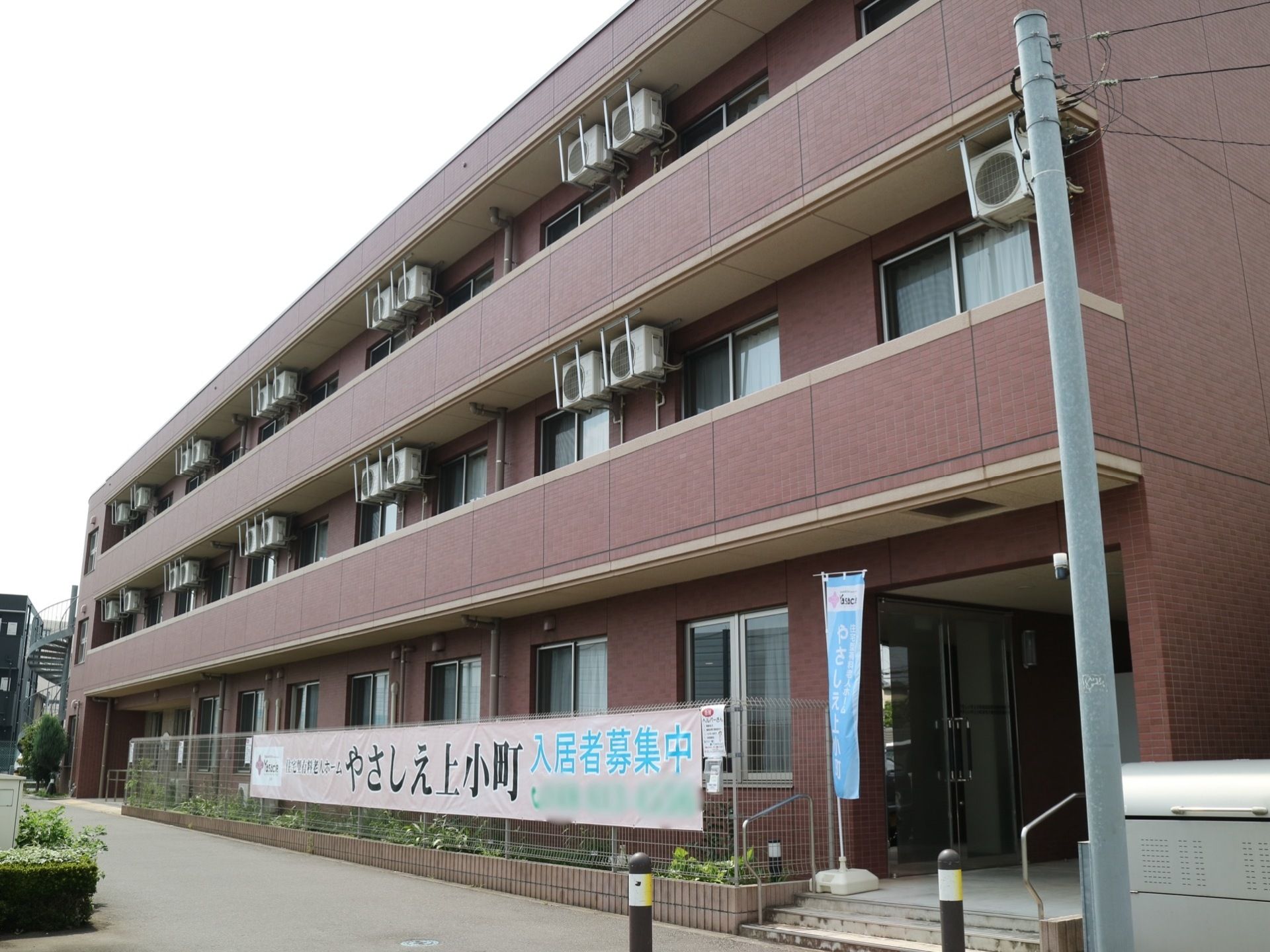 ブラウンを基調とした３階建てのグループホームである。２階以上の部分にはベランダが設けられており、エアコンの室外機が設置されている。