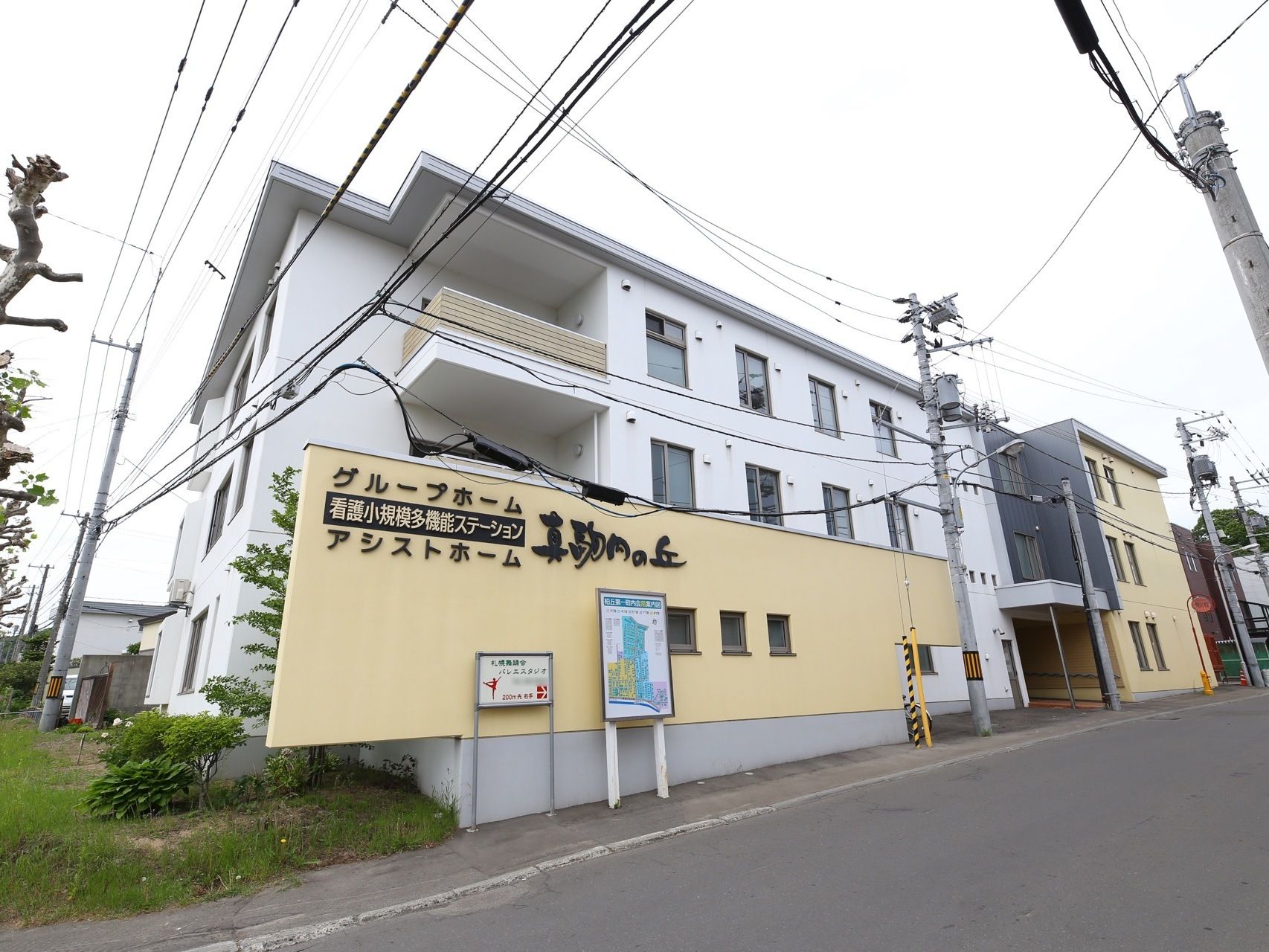 白、黒、ベージュを基調とした３階建ての施設。施設名が書かれた塀の前に、施設内の案内板が掲示されている。