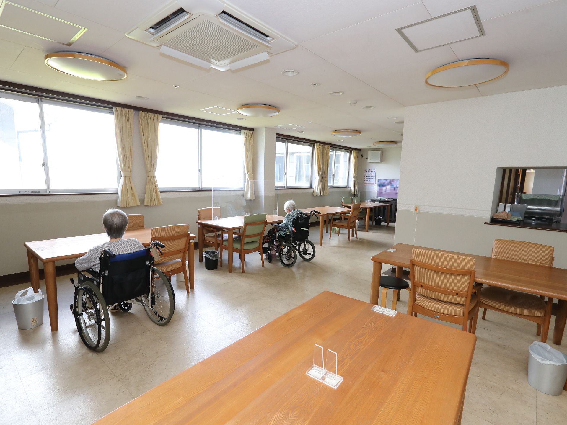 壁１面が窓ガラスになっている広間に並んだ木製の机近くに座る入居者様