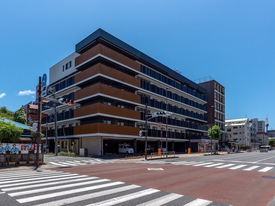 6階建ての建物はグレーとブラウンの外壁で各フロアにベランダが付いている。そばには交差点があり、建物も多い。