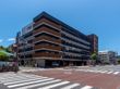 6階建ての建物はグレーとブラウンの外壁で各フロアにベランダが付いている。そばには交差点があり、建物も多い。
