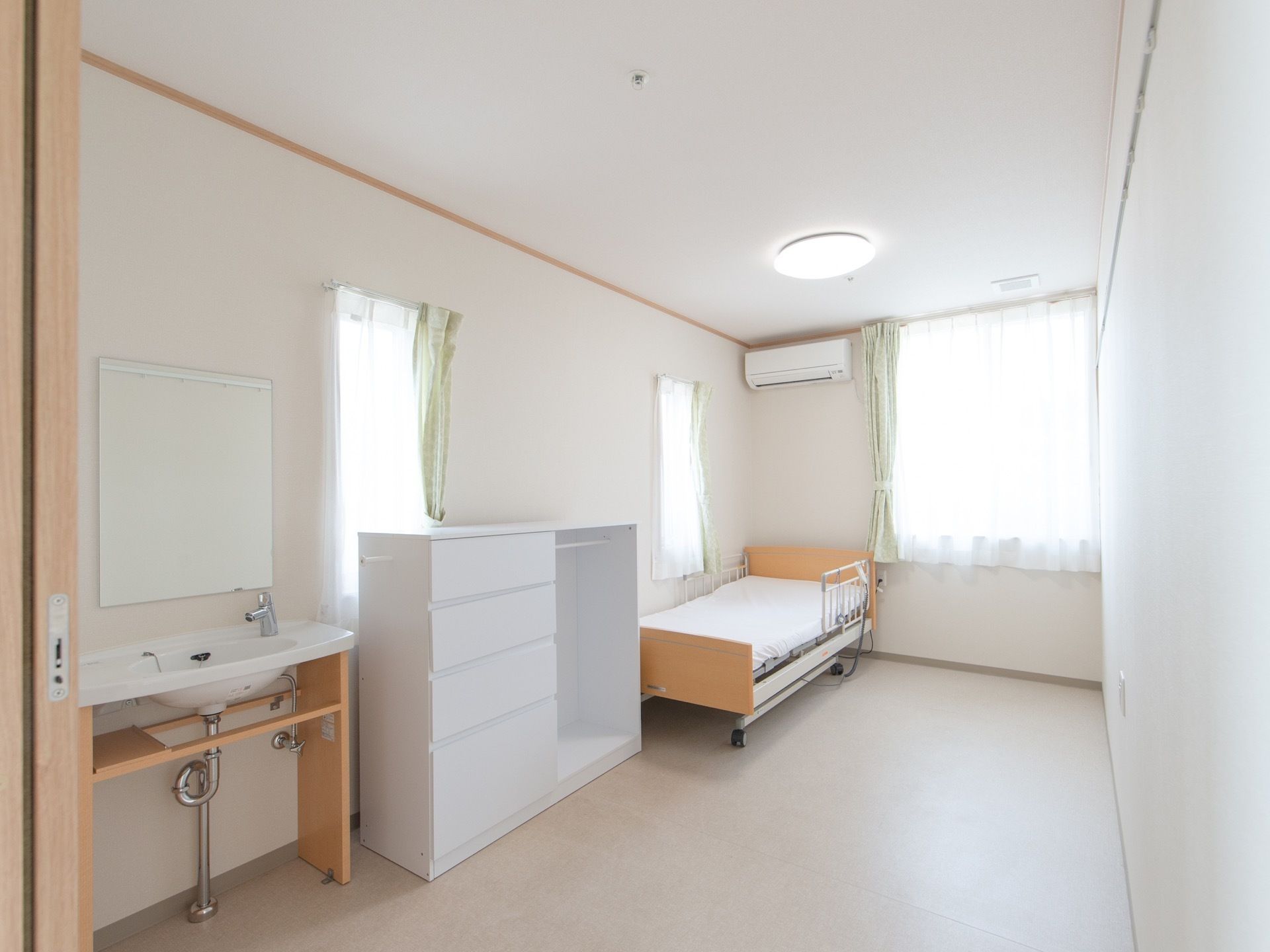 部屋の入り口から左側の壁に沿って、洗面台、収納棚、ベッドの順に置かれている。ベッドの上の壁にはエアコンが設置されている。
