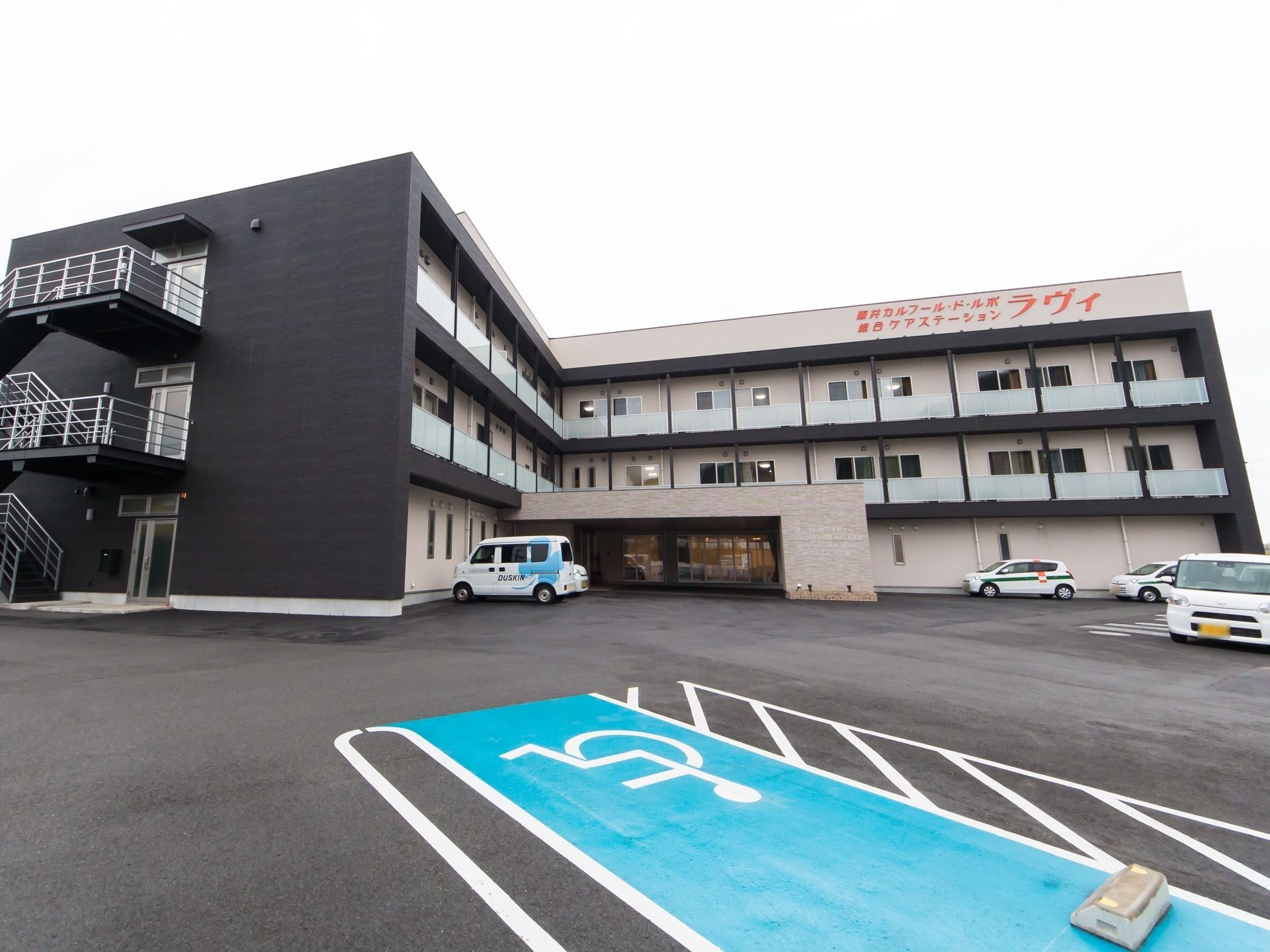白色と黒色を基調とした外壁を持つ3階建ての施設である。車椅子のマークが描かれた駐車スペースが水色で塗られている。