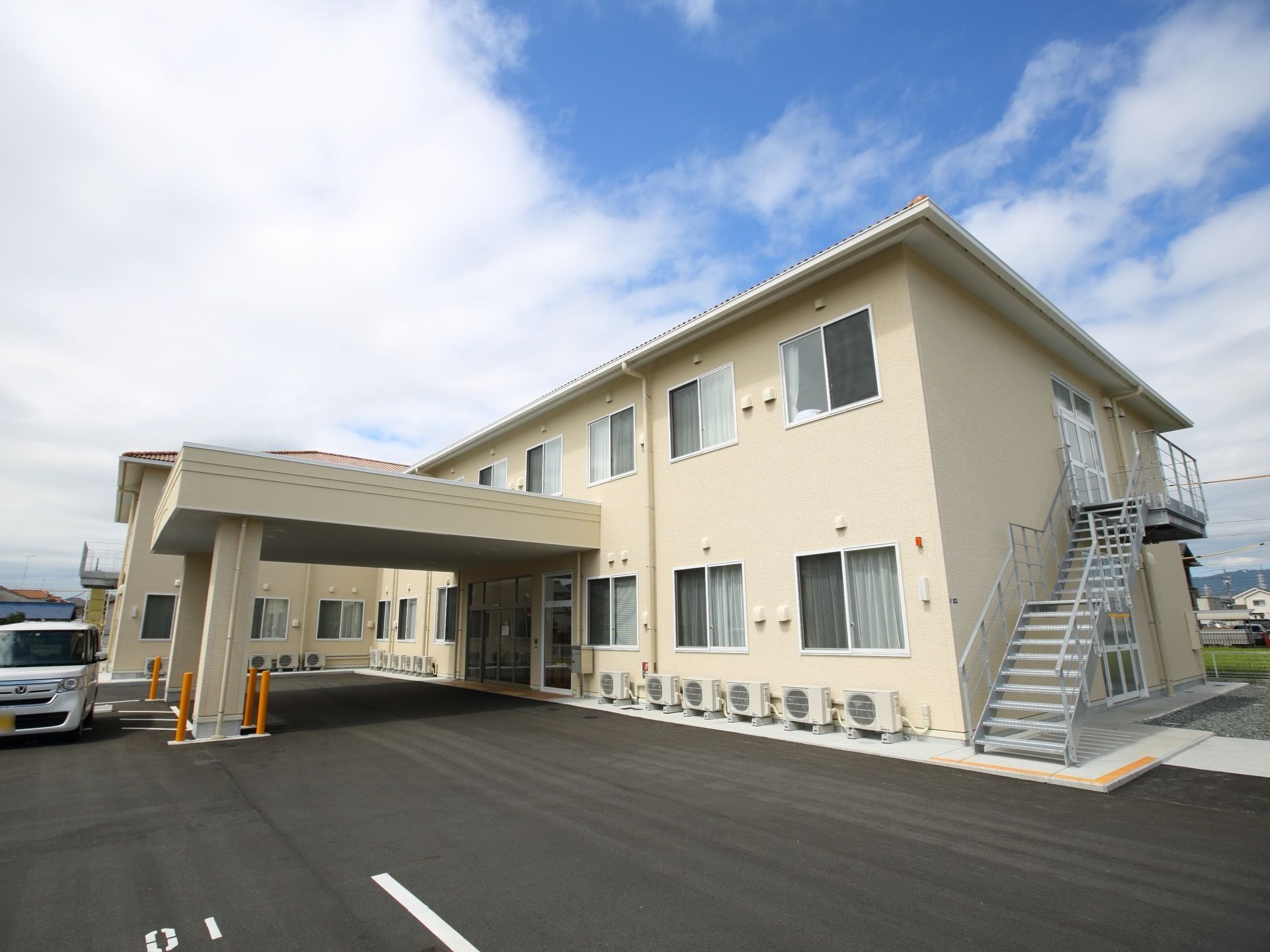 白を基調とした２階建てのグループホームで、建物横には非常階段がある。外壁沿いには、エアコンの室外機が複数置かれている。