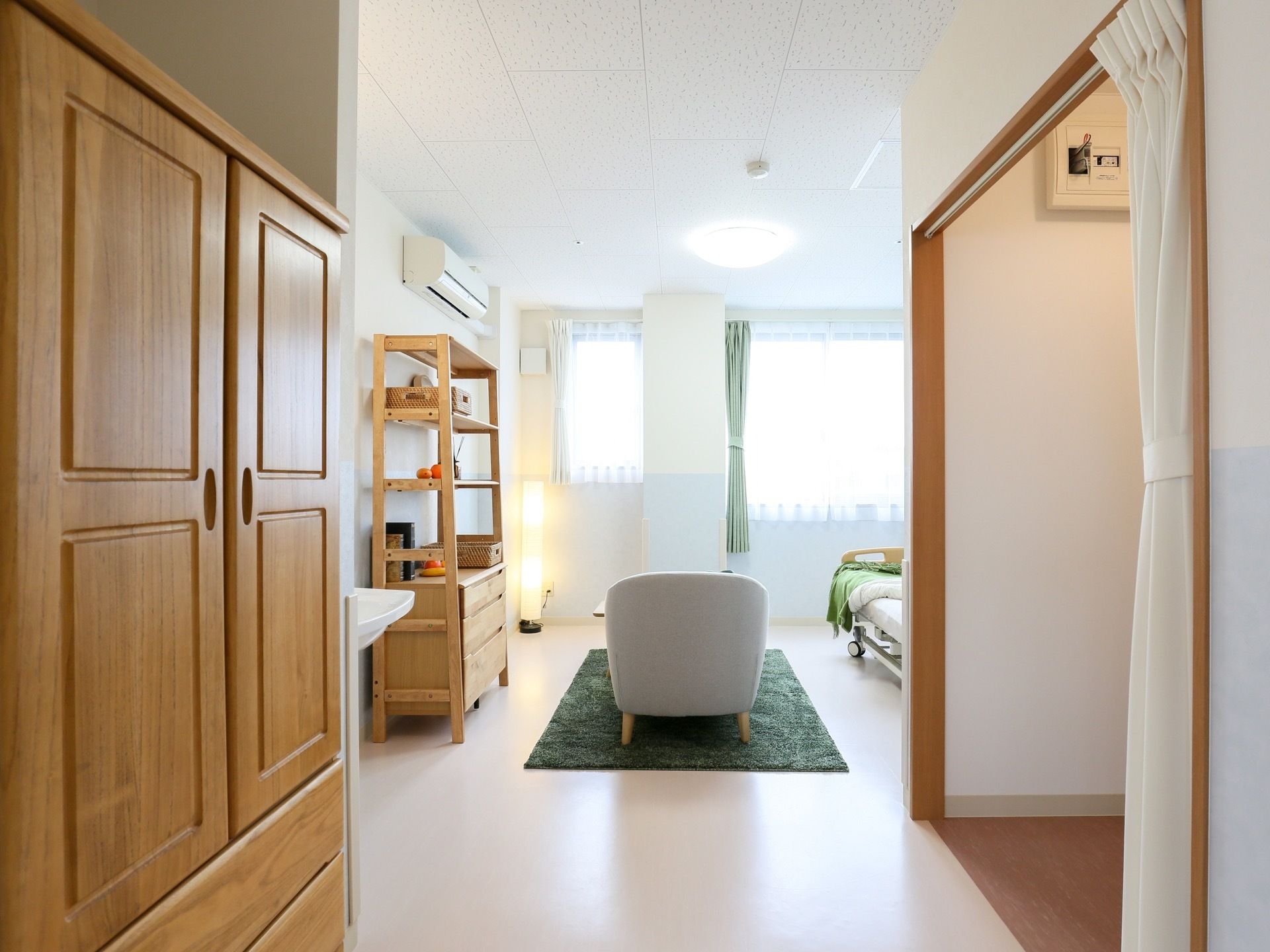 部屋の中央にカーペットが敷かれ、座椅子がある。木製の収納家具があり、座椅子横の壁沿いに整理棚がある。壁にエアコンがある。
