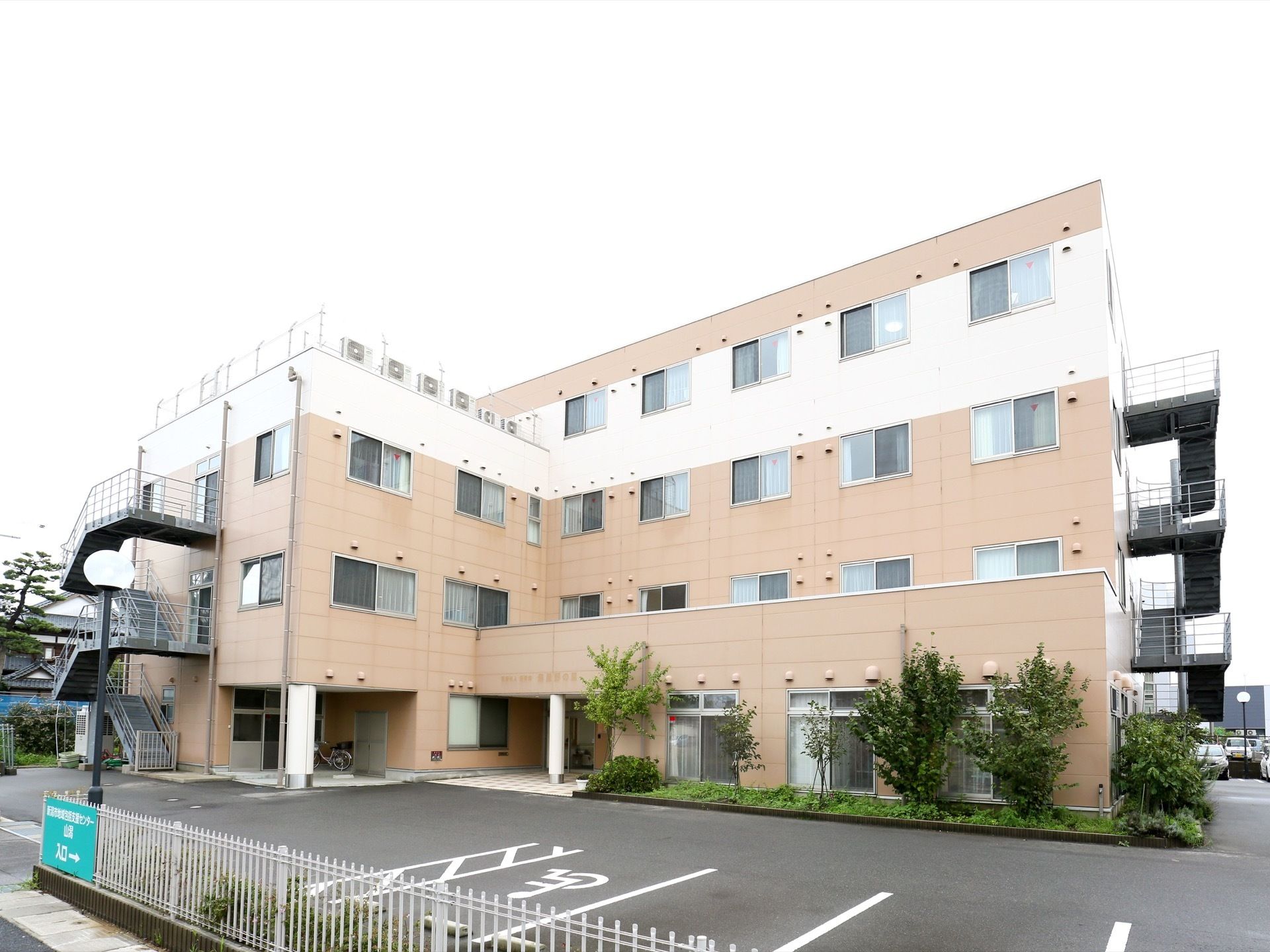 施設は鉄骨造りの４階建てである。外壁は、ベージュ系の色となっている。建物の両端に、外階段がある。建物の前面に、駐車場がある。