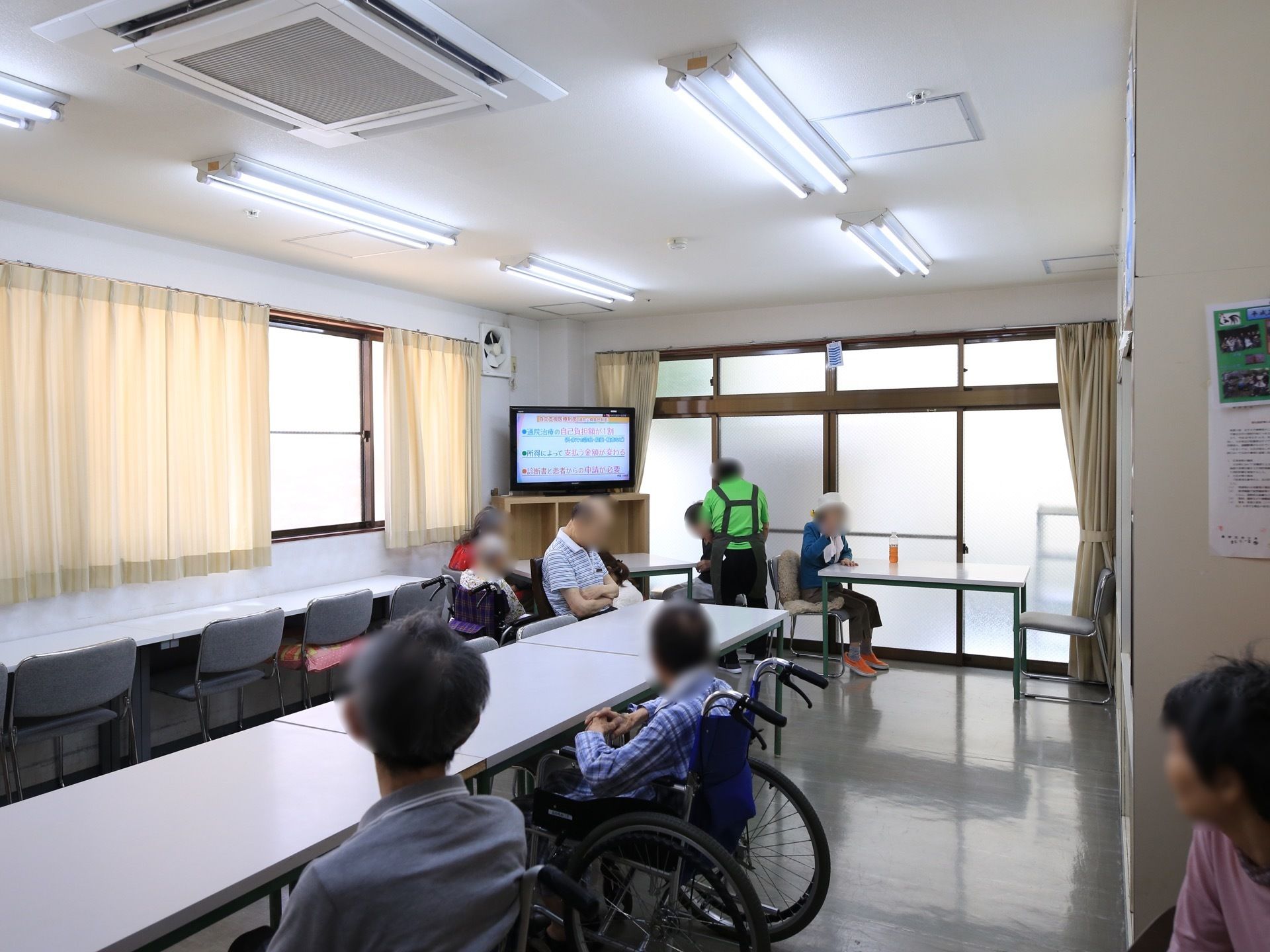 白い長机が並んだフロアで、入居者様とスタッフが集まっている。テレビを鑑賞したり、お茶を飲んだりしてくつろいでいる。