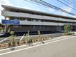 施設は３階建てで、白とグレーで統一された外壁となっている。正面の駐車場には、白い車がたくさん停められている。