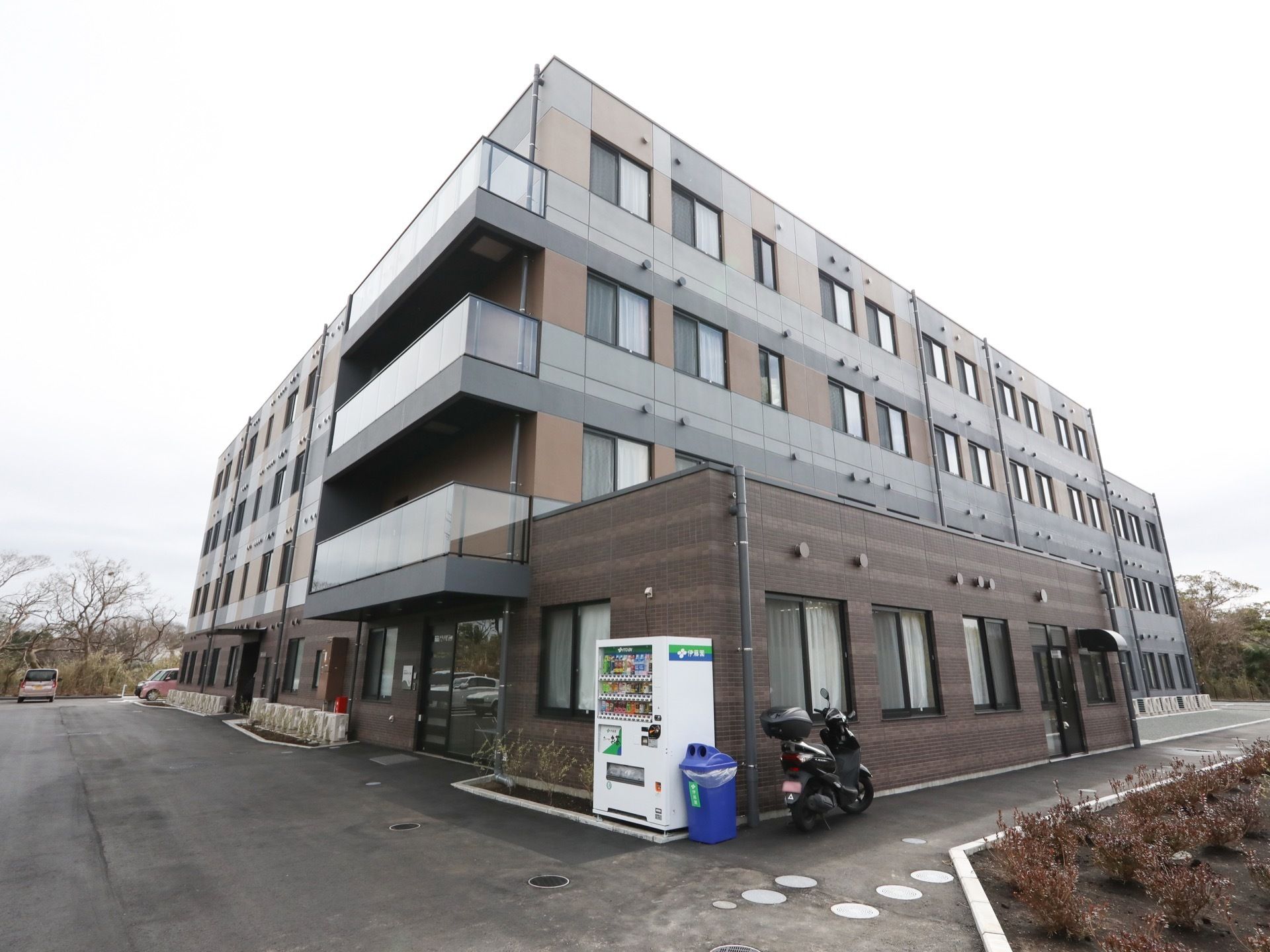 ４階建ての落ち着いたカラーで施された建物である。敷地内には駐車場が設けられており、車が停められている。