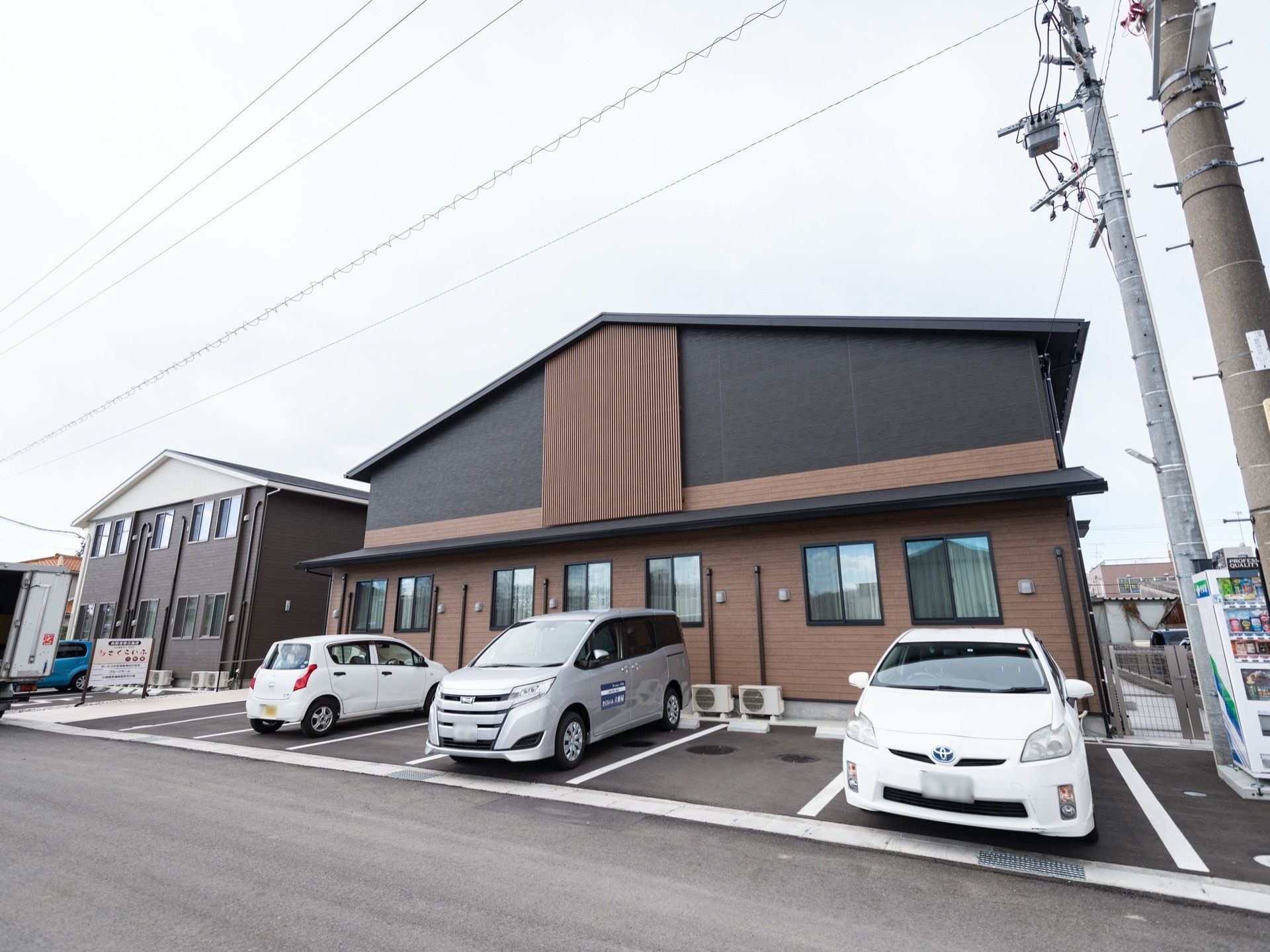 周囲の住宅街に馴染んだ、２階建て施設の建物の様子。車道脇に建っているため、車を横付けしやすい。施設の看板が見られる。